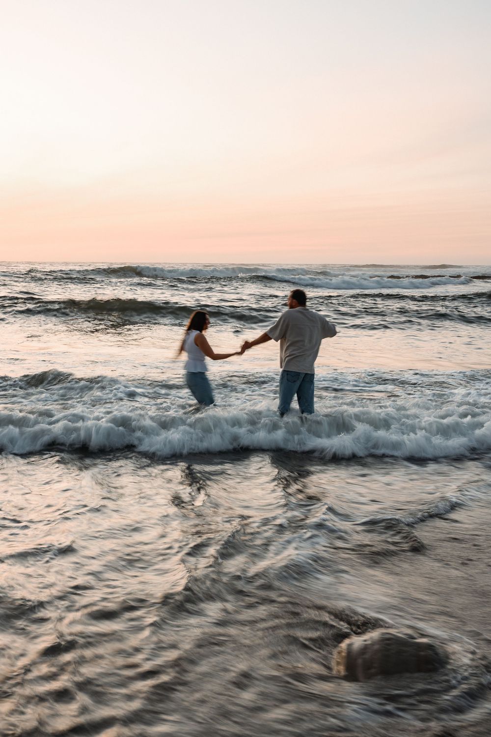 Couple holding hands standing in ocean waves at sunset on a sandy beach.