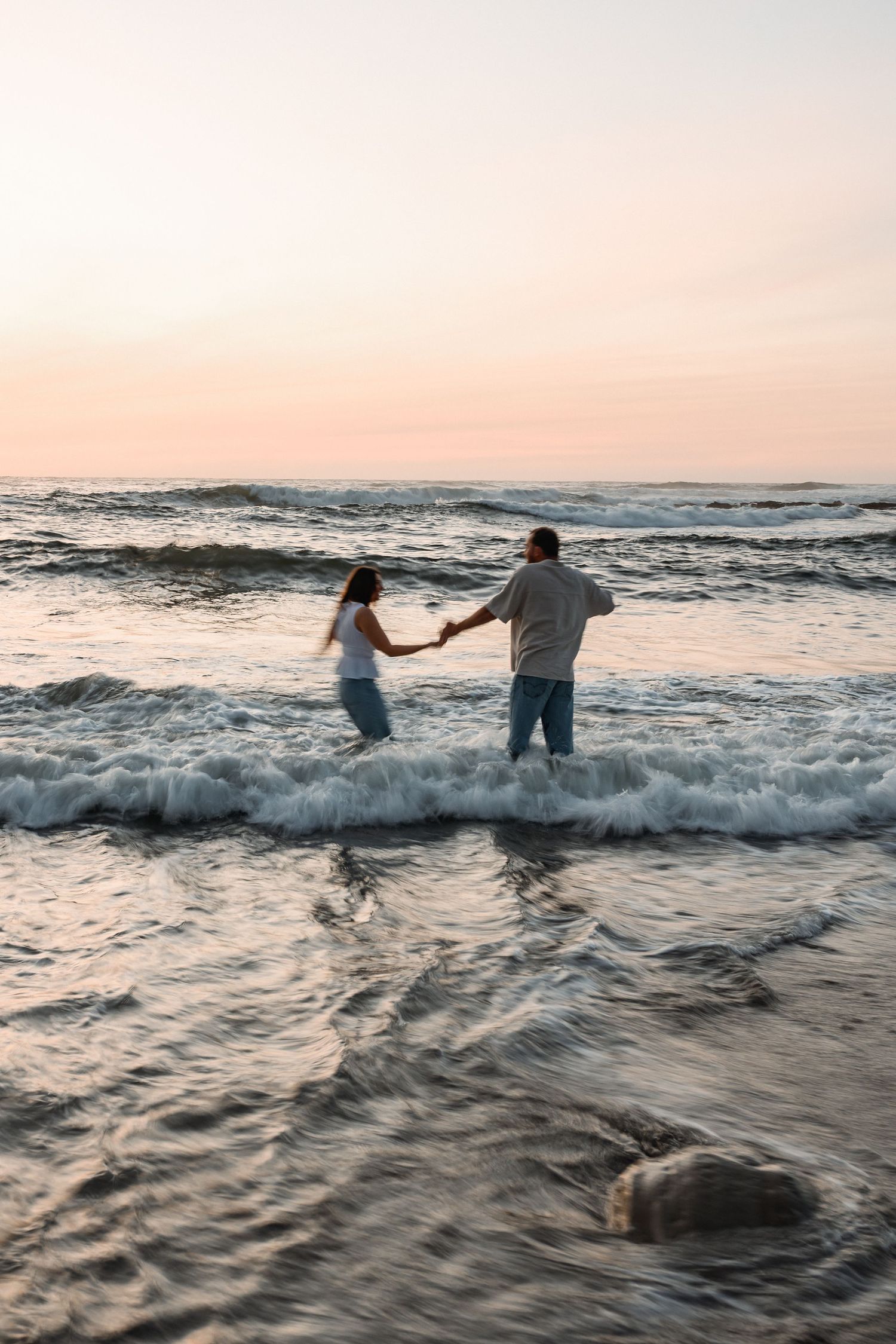 Couple holding hands standing in ocean waves at sunset on a sandy beach.
