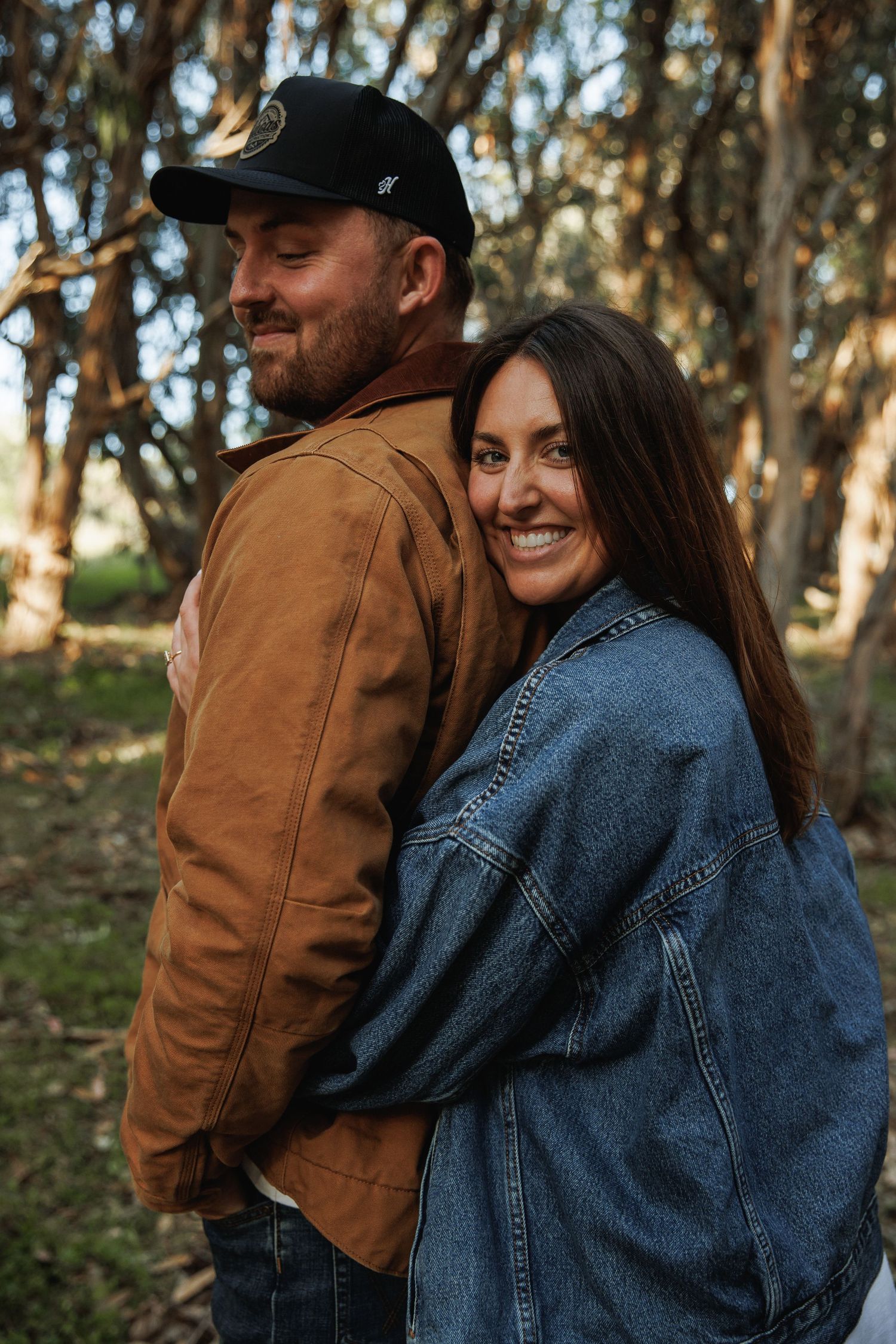 Woman smiling and hugging man from behind in a sunlit forest, both wearing casual jackets.