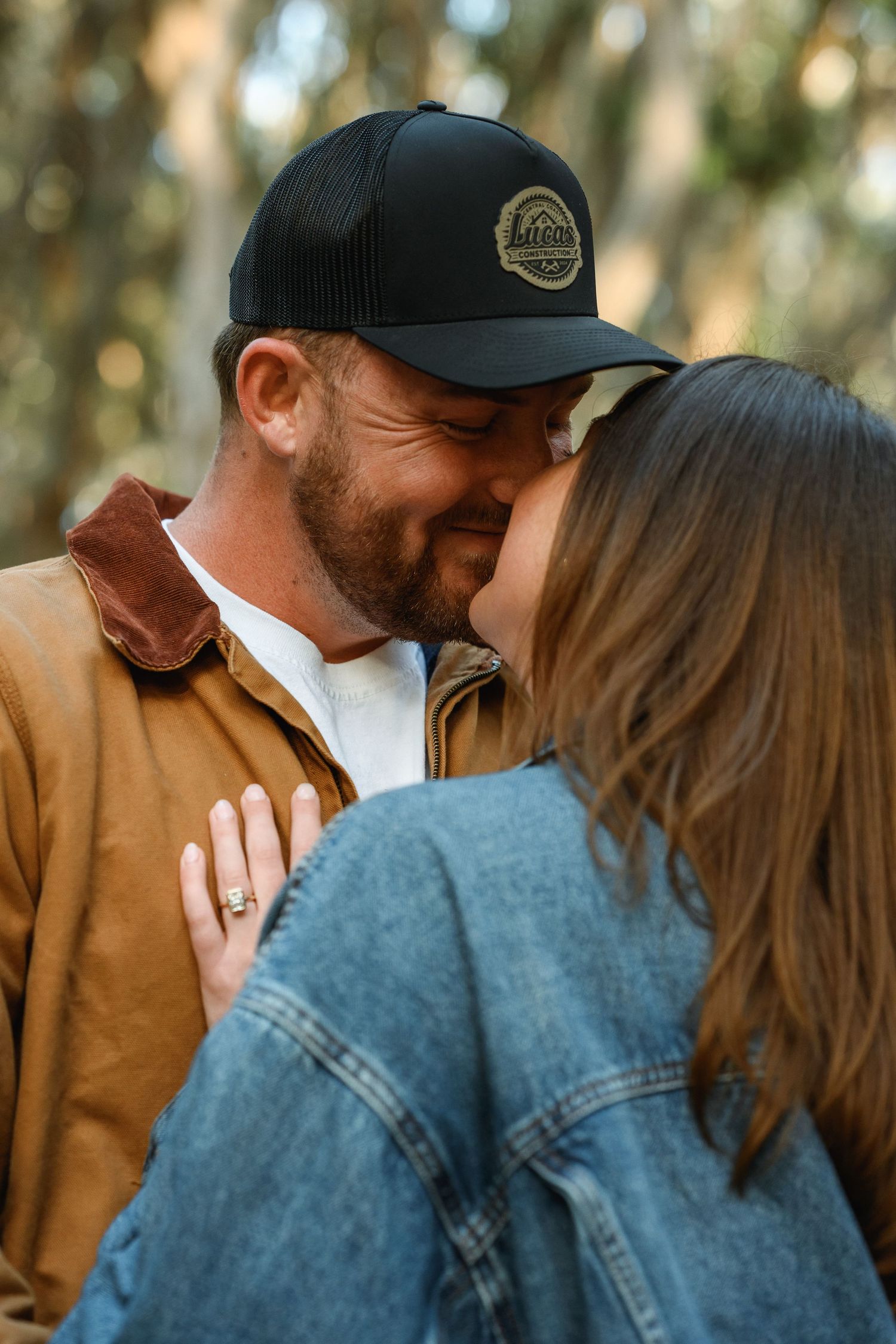 Close-up of couple almost kissing in a forest, man in brown jacket and black cap, woman showing ring.