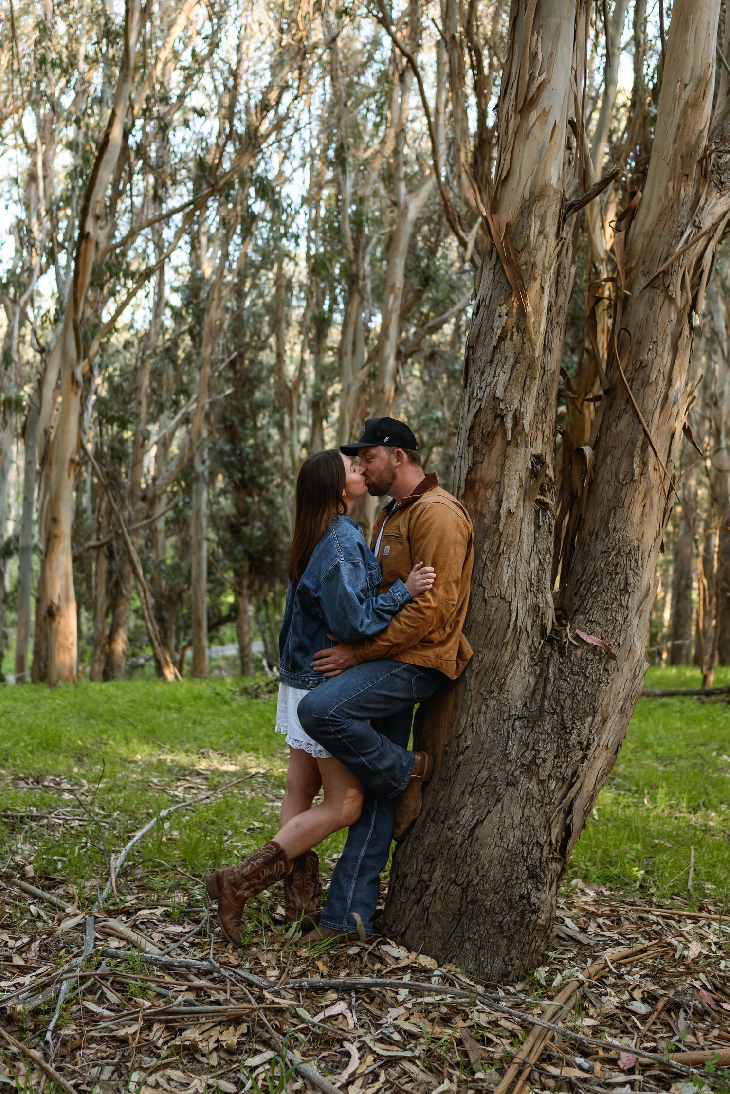 Couple kissing against a tree in a eucalyptus forest, woman in denim jacket and brown boots.