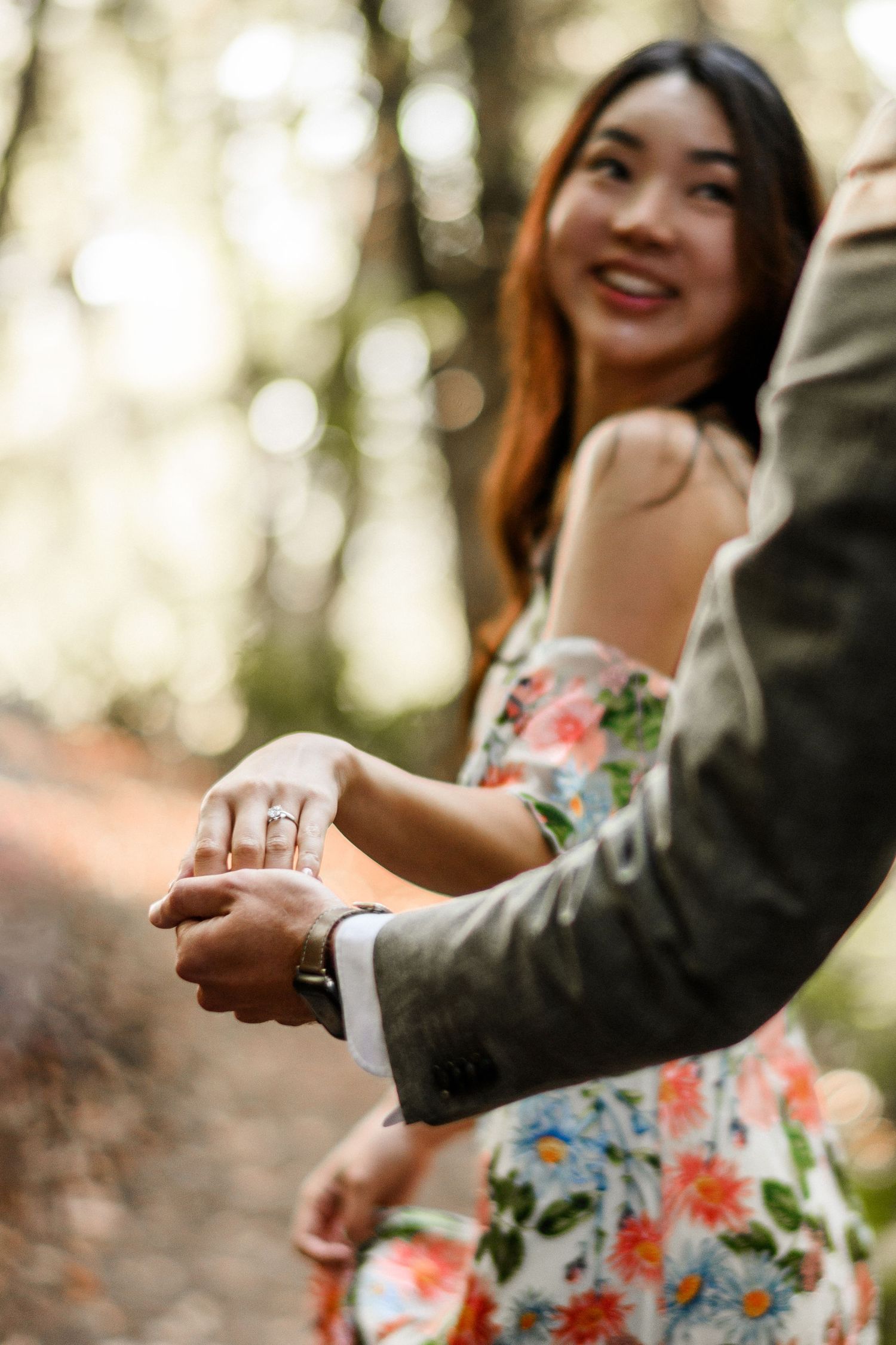 Close-up of engagement ring on woman's hand held by man in suit, smiling woman in background.