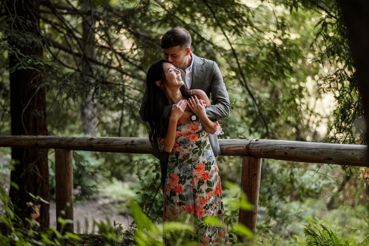 Couple sharing a tender kiss on wooden bridge surrounded by lush green redwood forest.