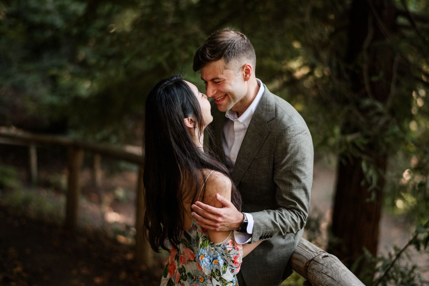 Smiling man in gray suit gazing at woman in floral dress near wooden fence in forest.