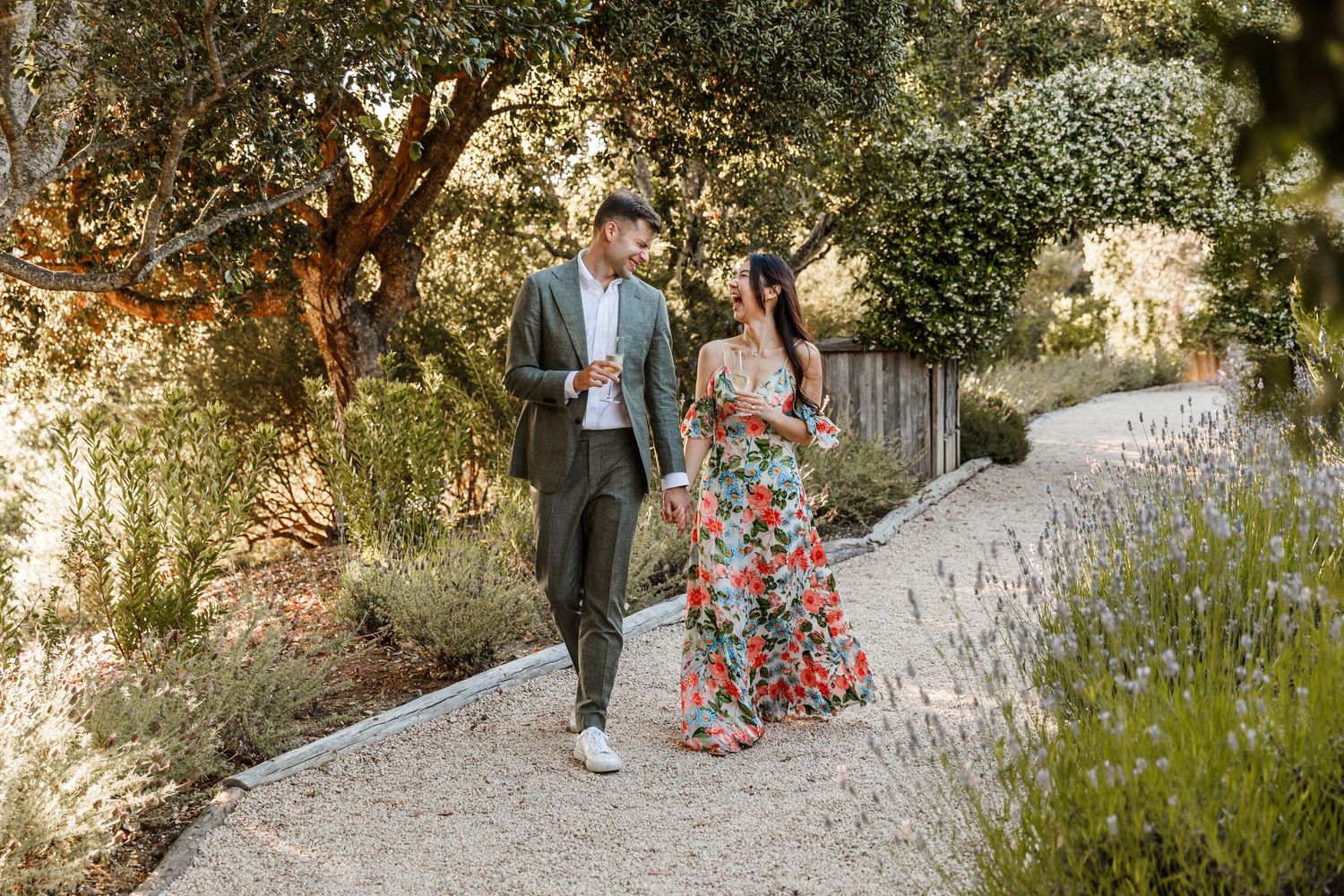 Elegant couple walking hand in hand on garden path lined with lavender and lush greenery.