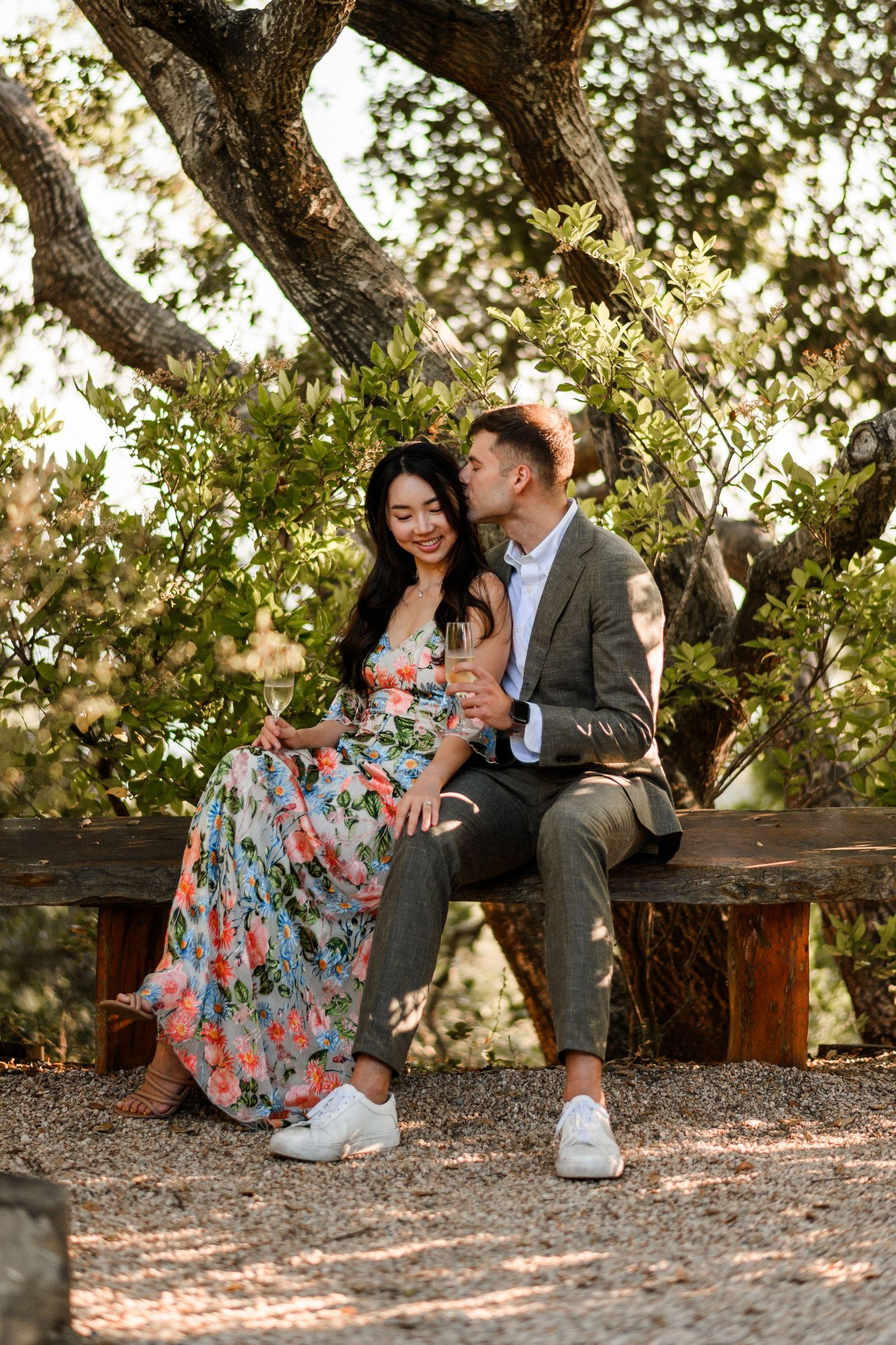 Couple sitting on rustic wooden bench under oak tree, man kissing woman in floral maxi dress.