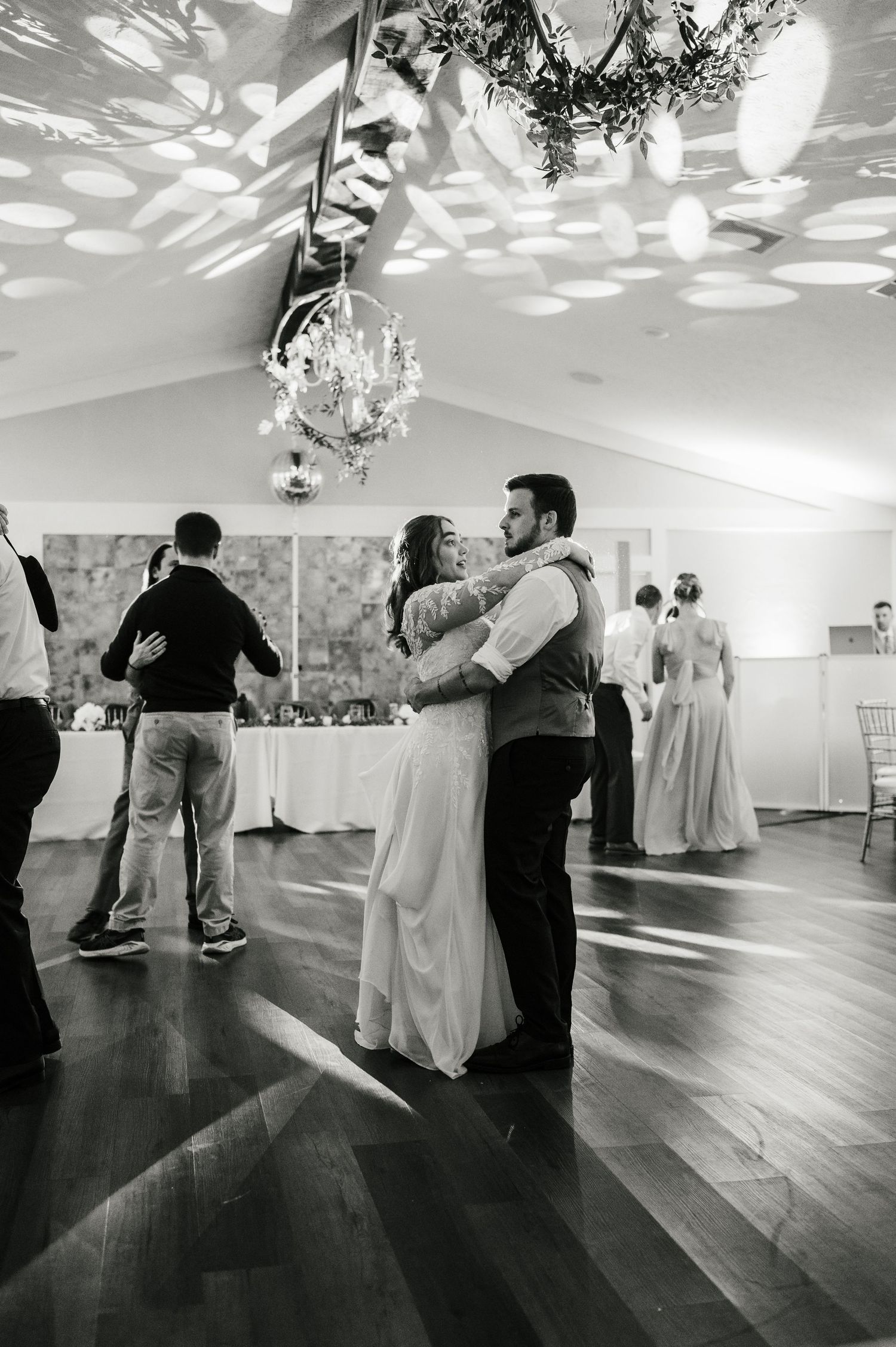 A couple slow dances under elegant chandeliers at a wedding reception with decorative light patterns on the ceiling.