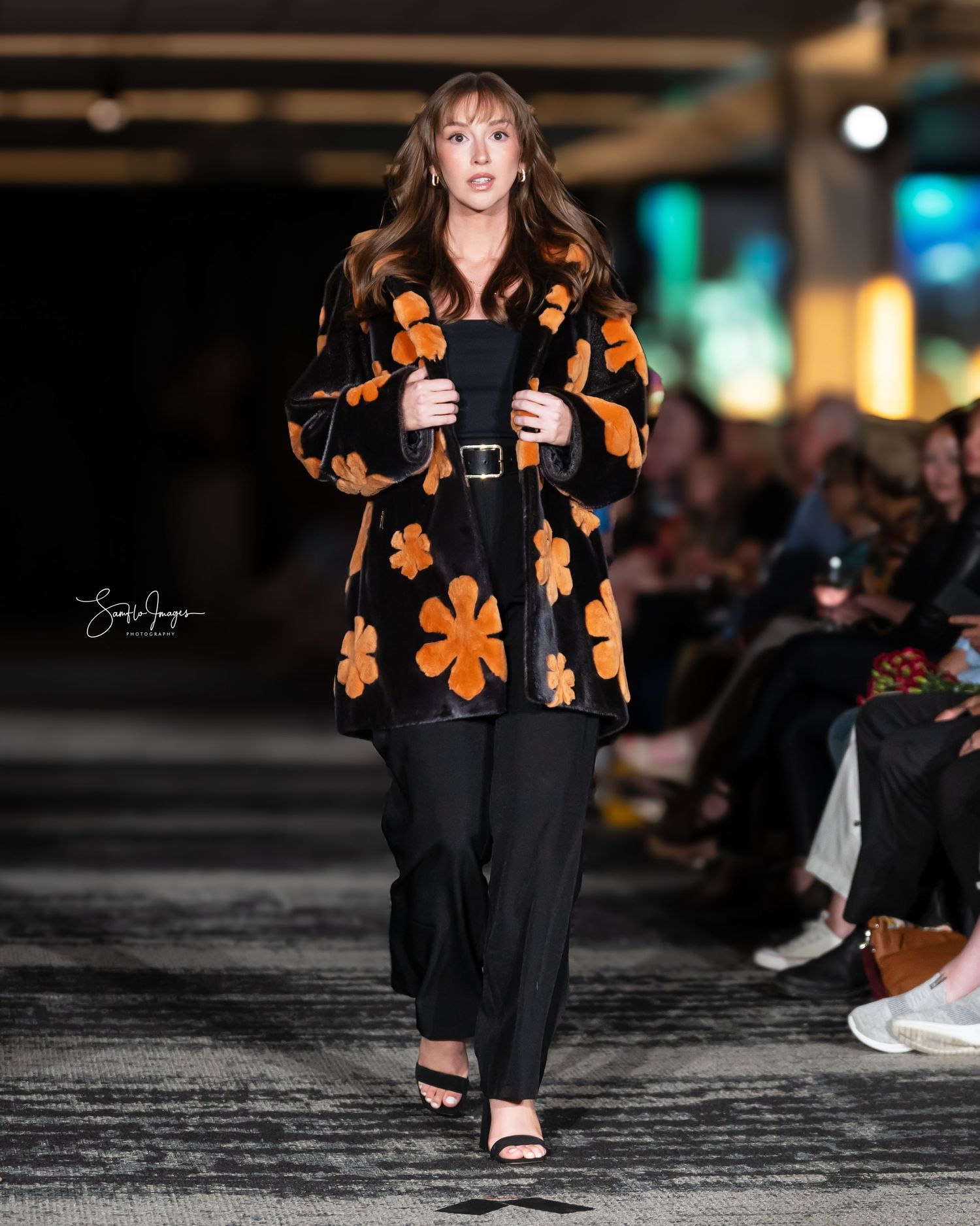 Model walking runway in black coat with orange flower pattern and black pants during fashion show.