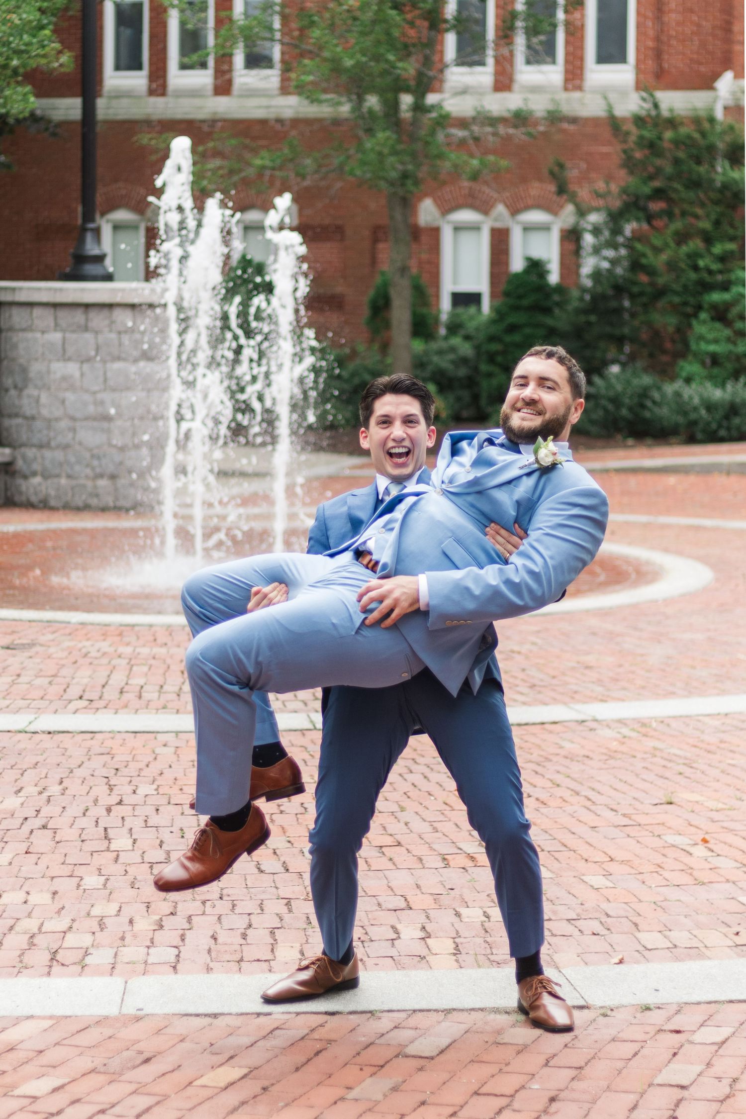 Playful groomsmen in blue suits pose by fountain on brick courtyard.