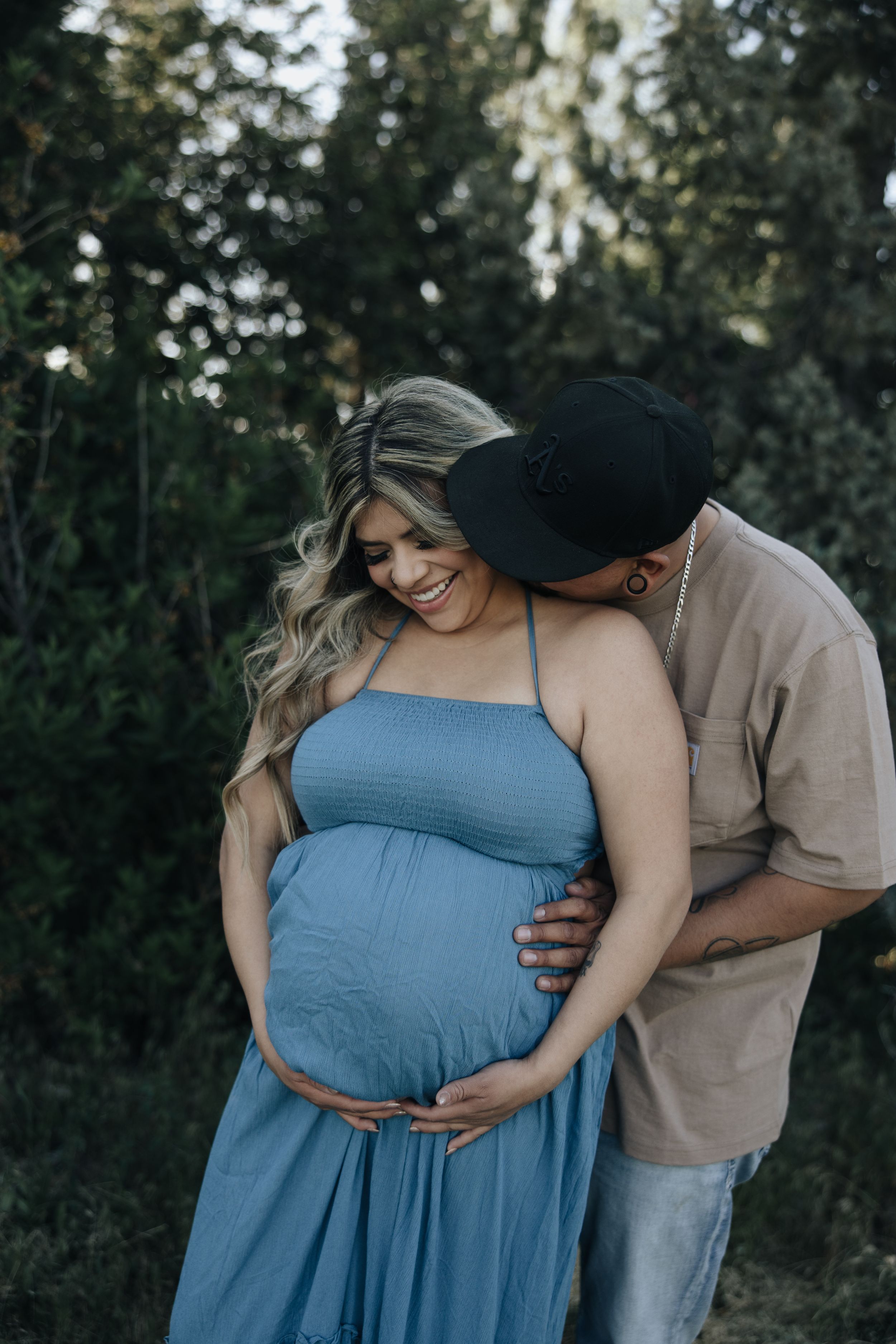 Intimate maternity portrait showing couple embracing outdoors in blue dress and casual wear against forest backdrop.