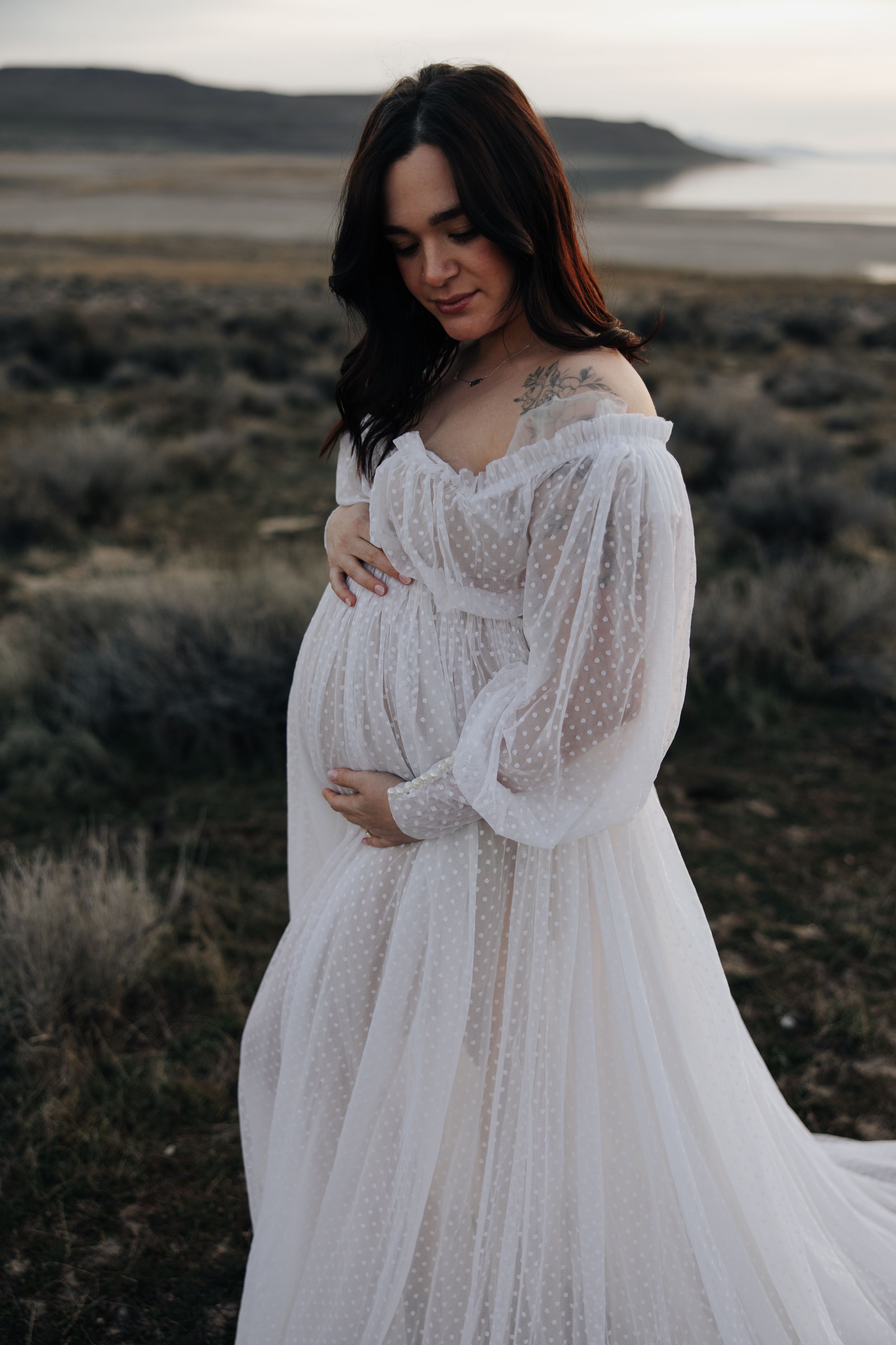 Romantic maternity portrait in white lace gown against rugged coastal backdrop.