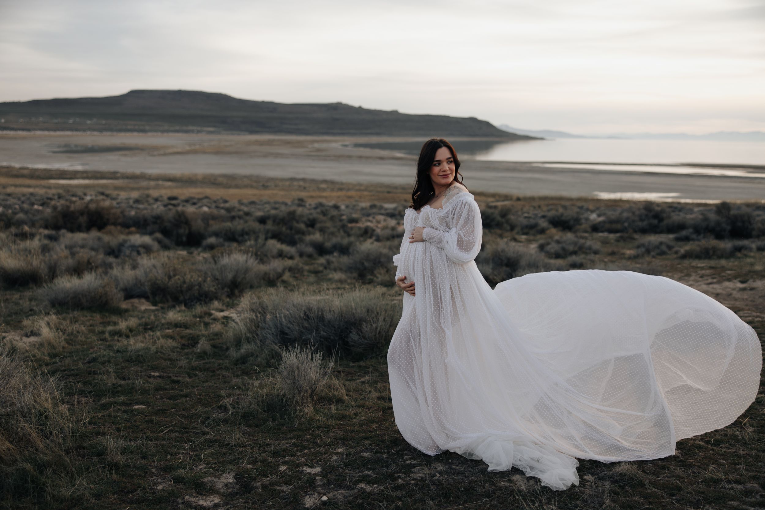 Ethereal maternity photo of woman in flowing white dress against coastal landscape.