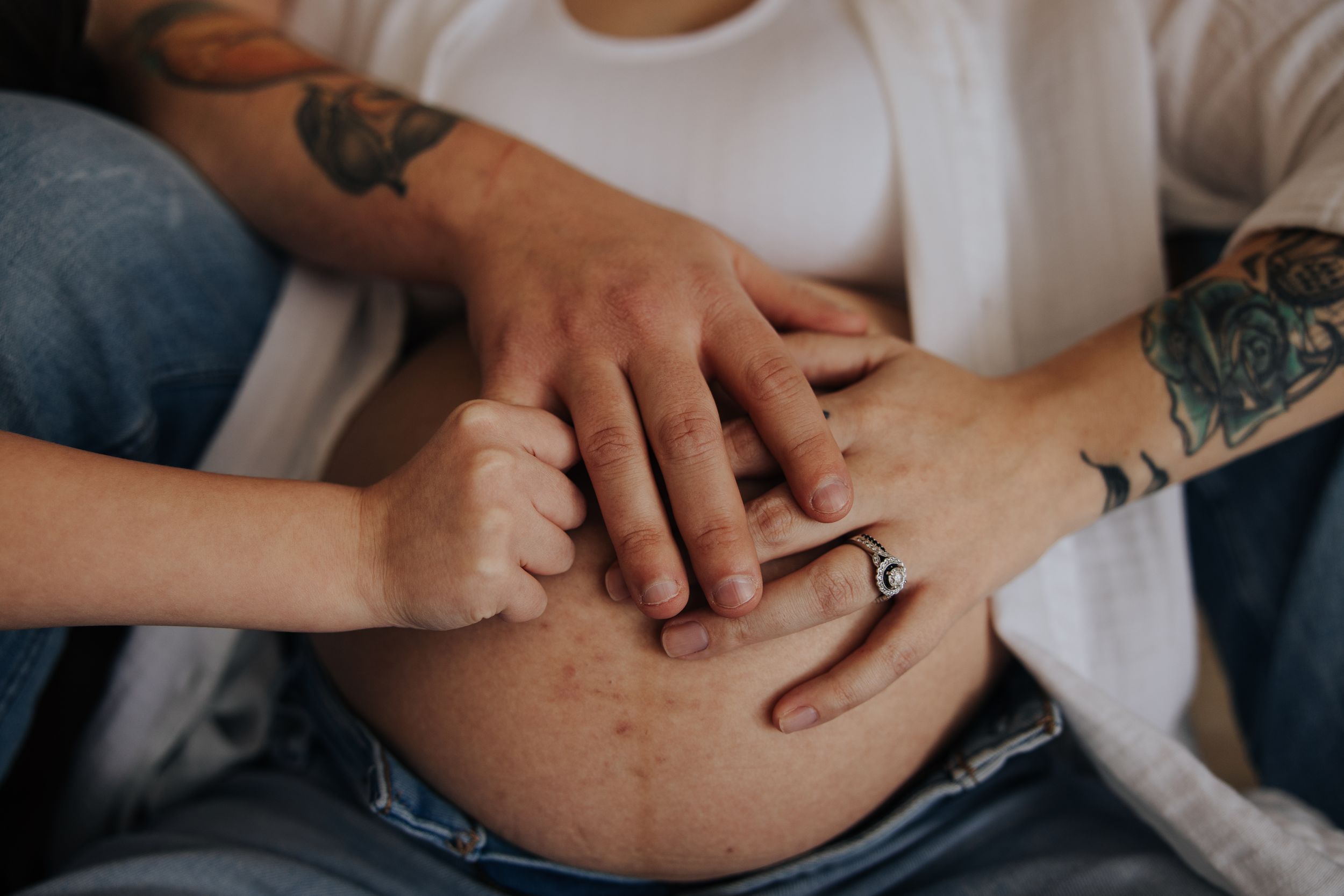 Close-up of hands resting on pregnant belly with tattoos and engagement ring.