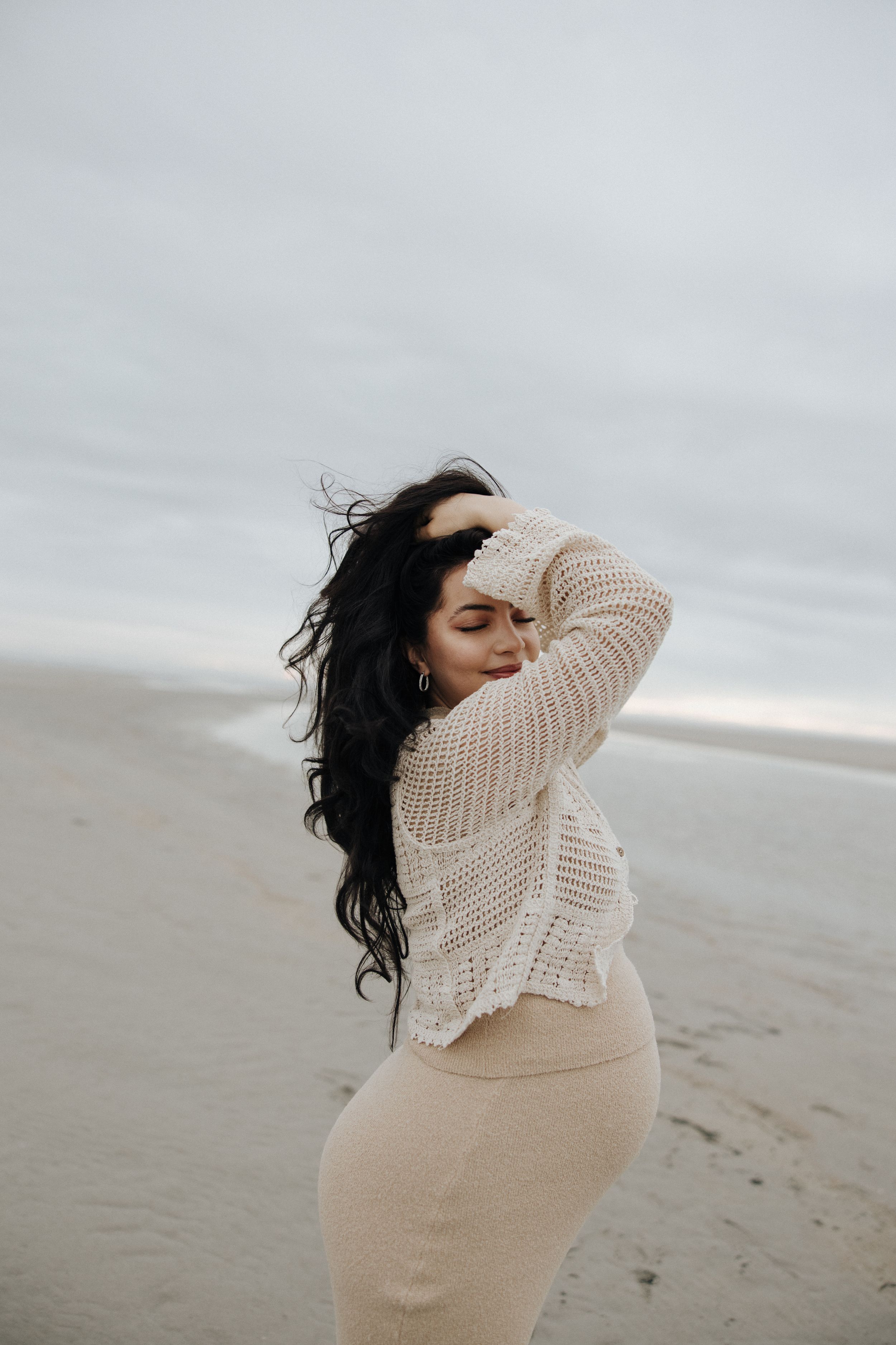A person in a cream sweater stands on a misty beach, their dark hair blowing in the wind.