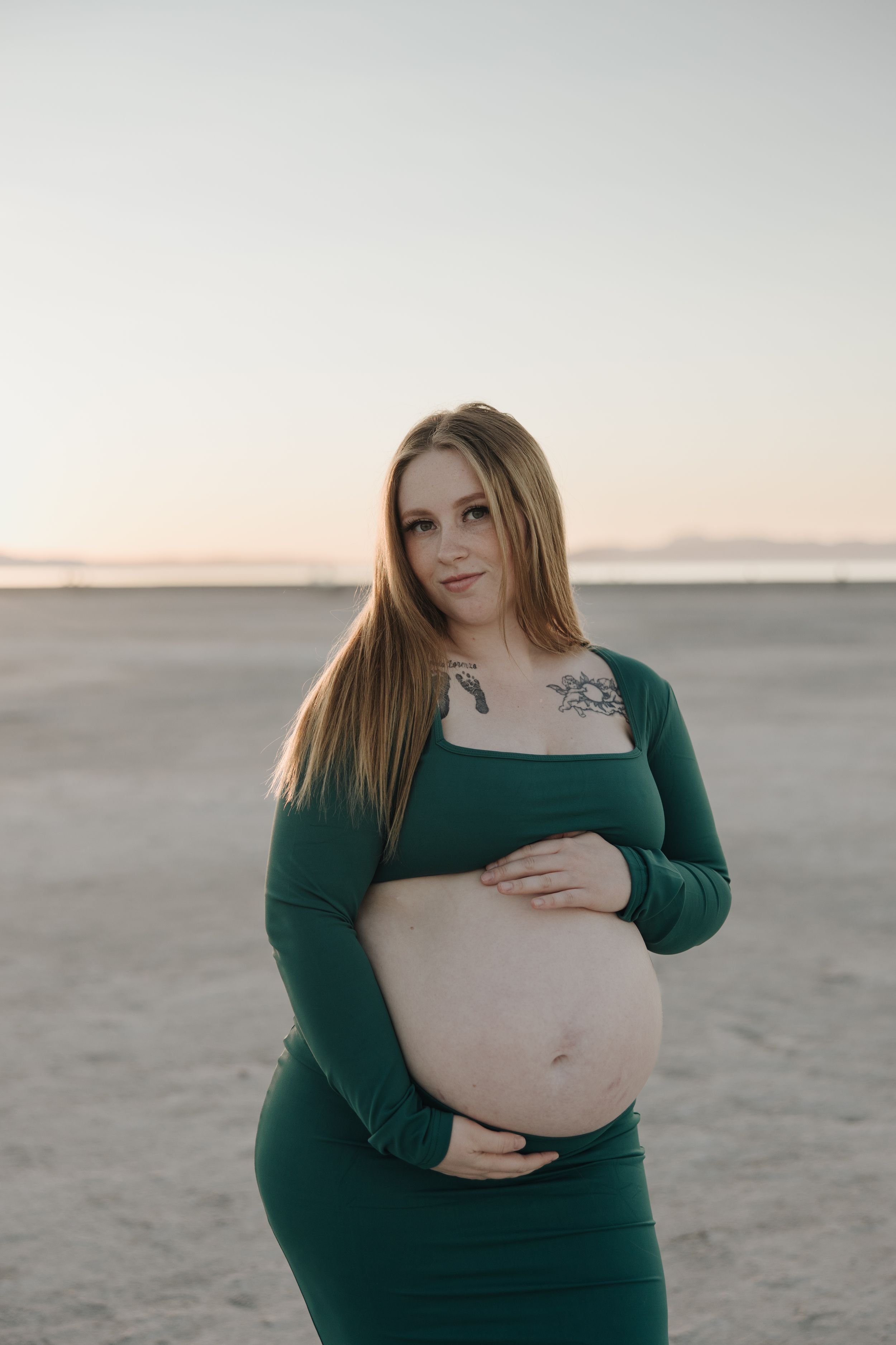 A pregnant woman wearing a green dress stands on a beach at sunset, cradling her baby bump.