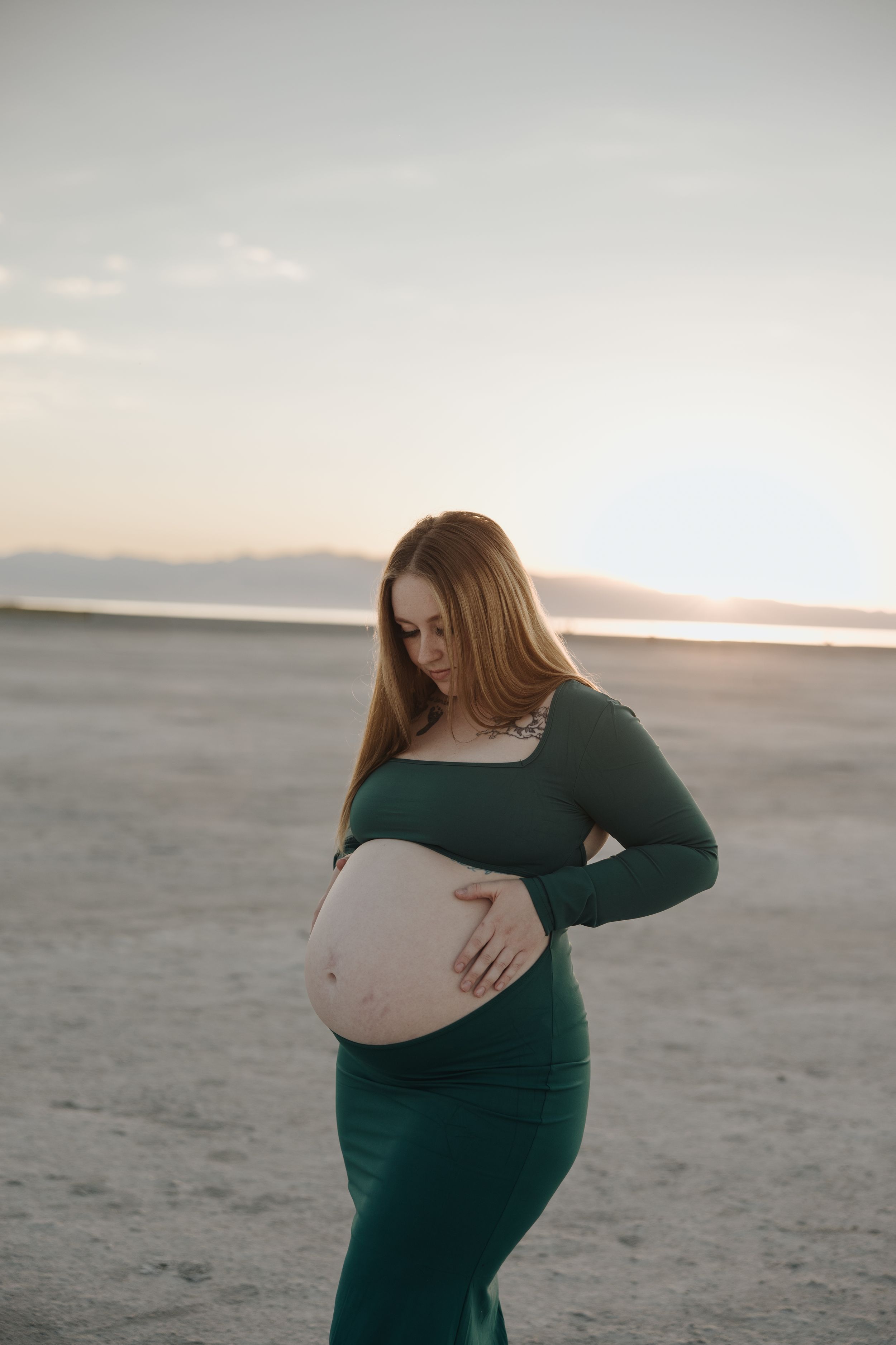 Expectant mother in emerald green dress stands on desert plain at sunset cradling her baby bump.