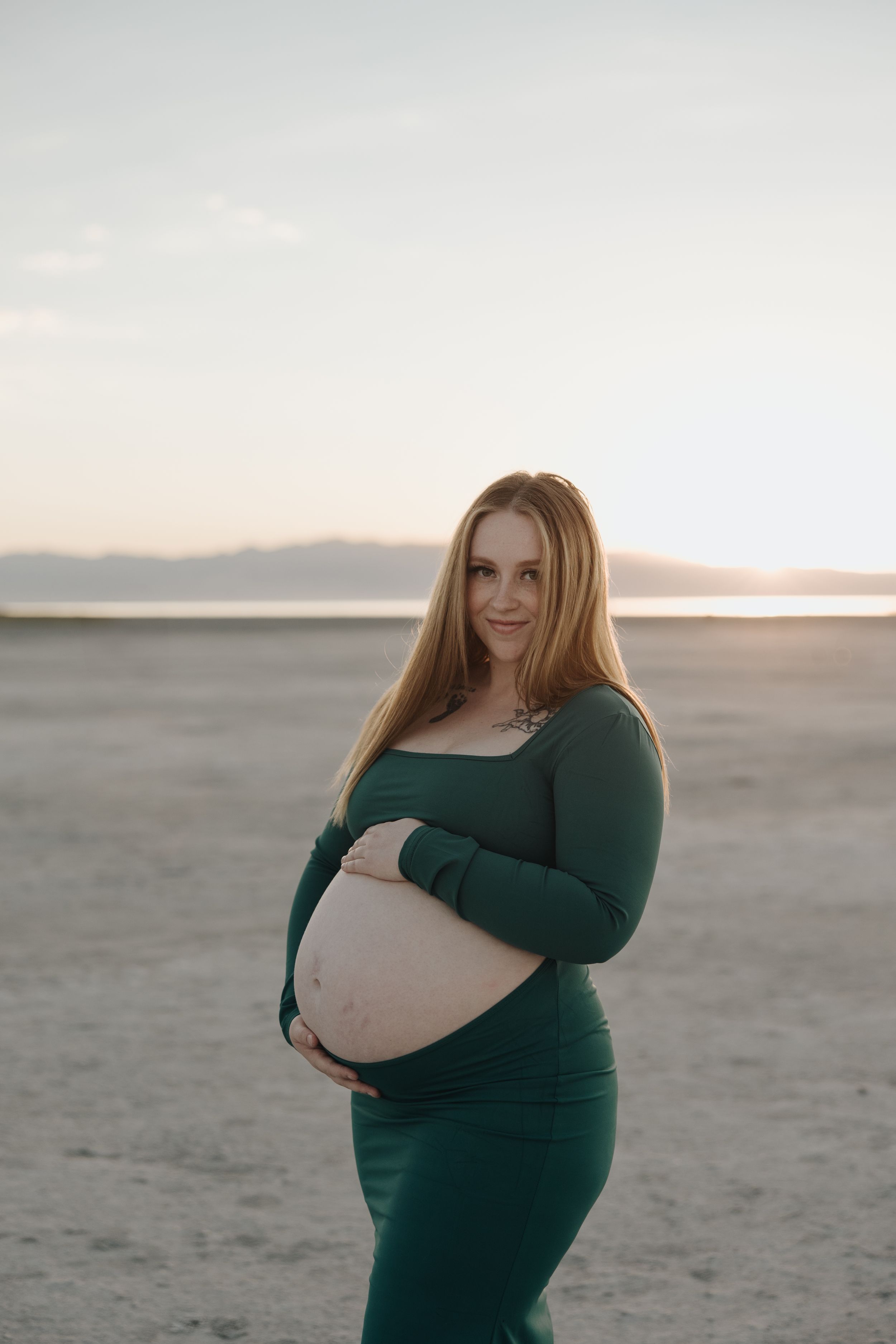 An expectant mother in a green dress stands on a desert landscape at sunset.
