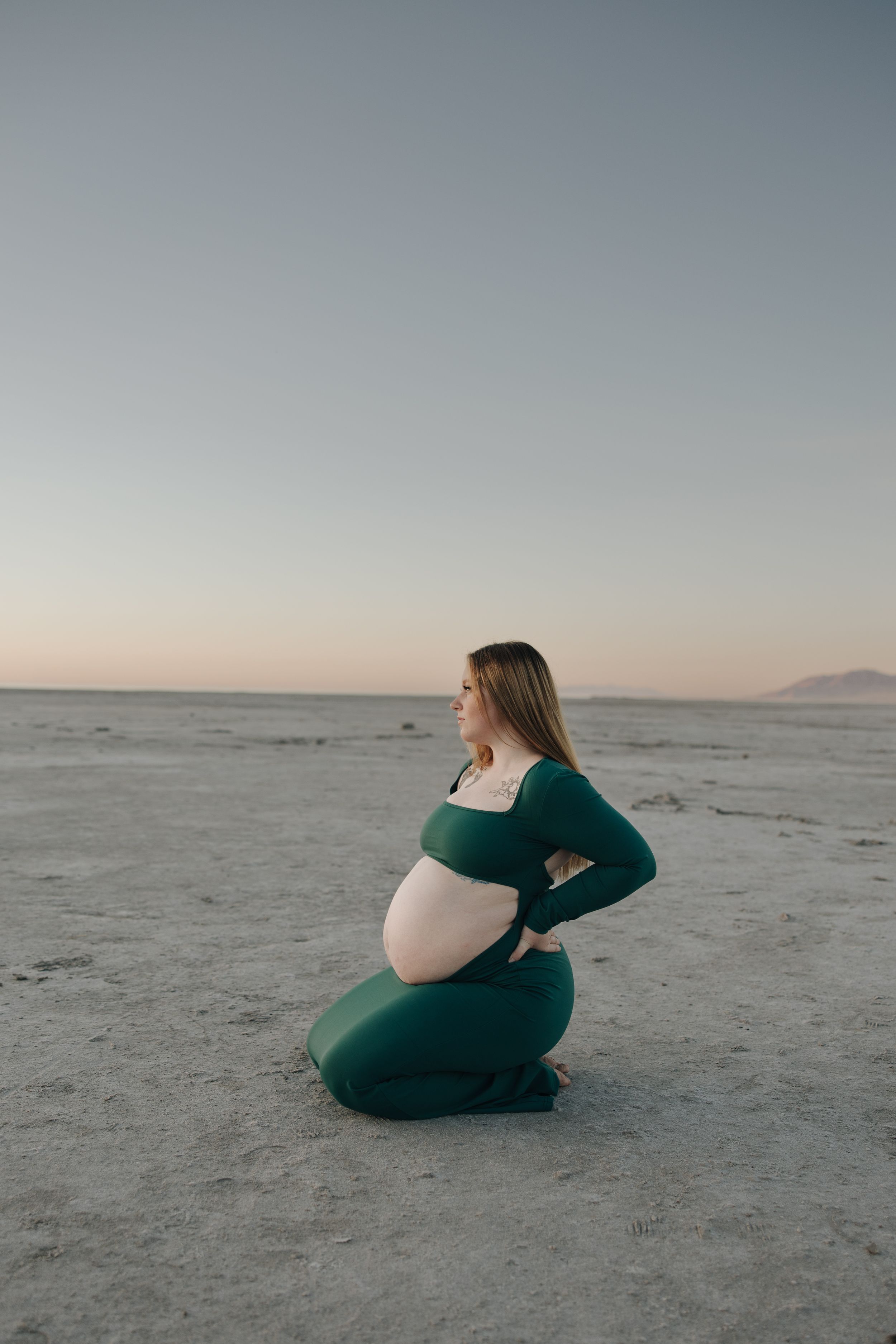 A pregnant woman in a green dress kneels thoughtfully on a vast, empty salt flat at sunset.