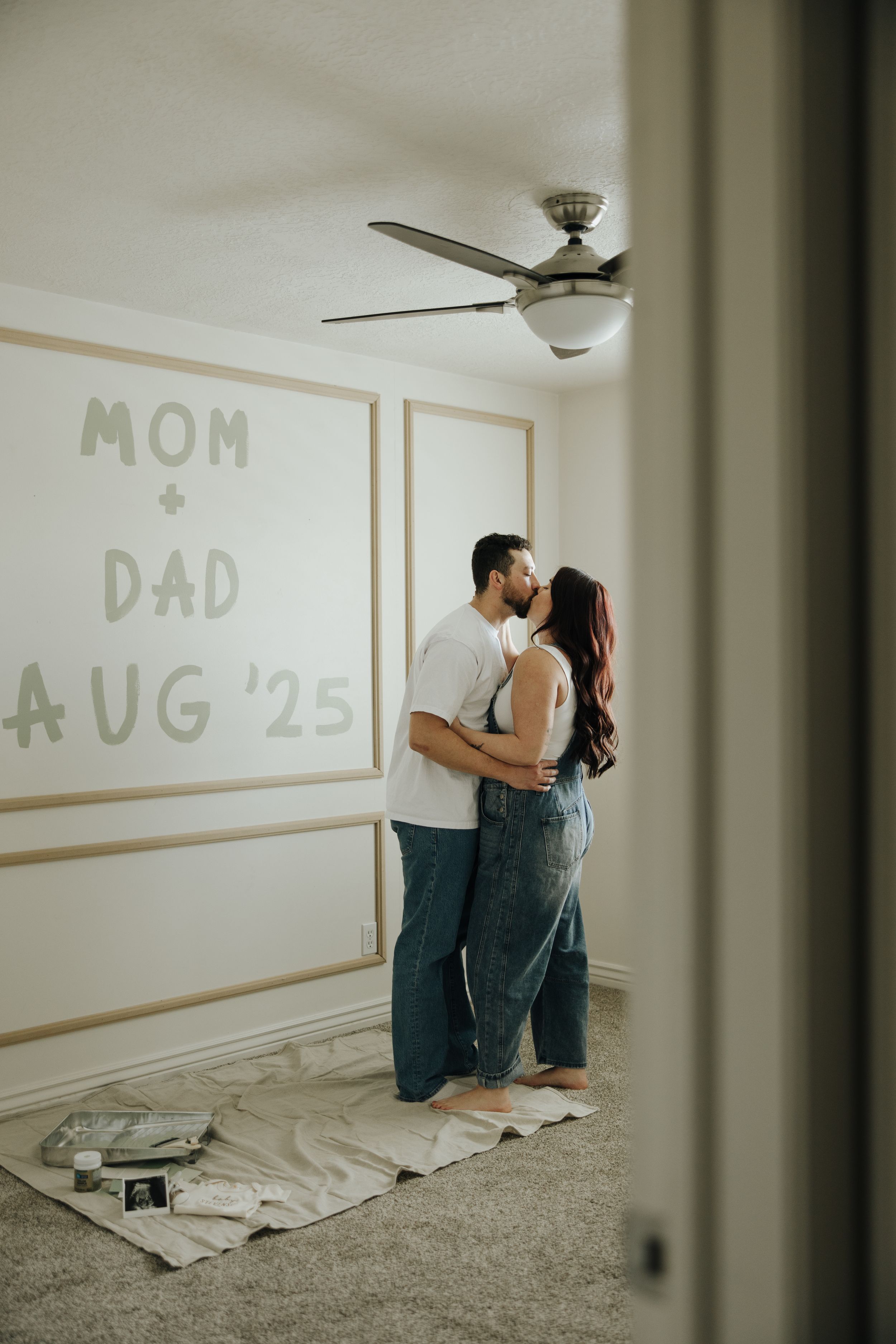 A couple embraces in an empty room with 'MOM + DAD AUG '25' written on the wall and a ceiling fan above.