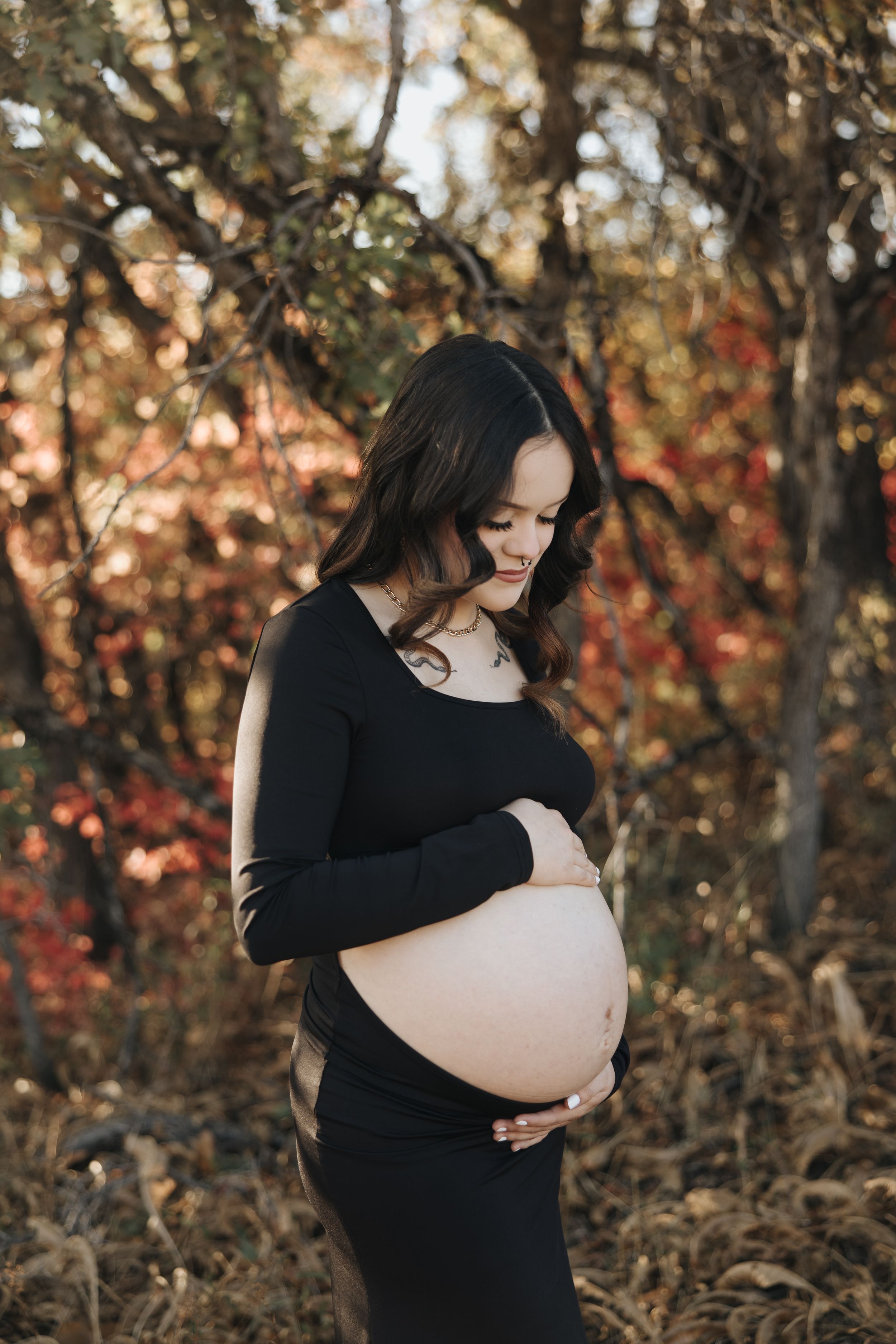 A pregnant woman in a black dress cradles her belly during an outdoor maternity photoshoot with autumn foliage in background.