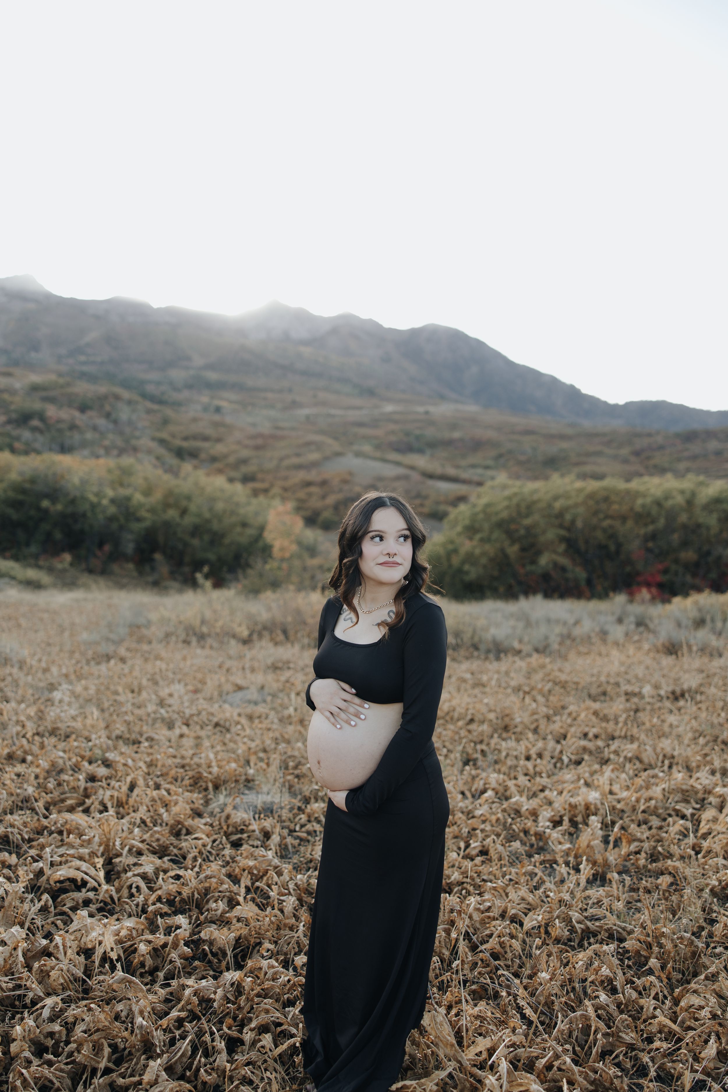 Expecting mother in long black dress stands in field with mountains in background during golden hour.