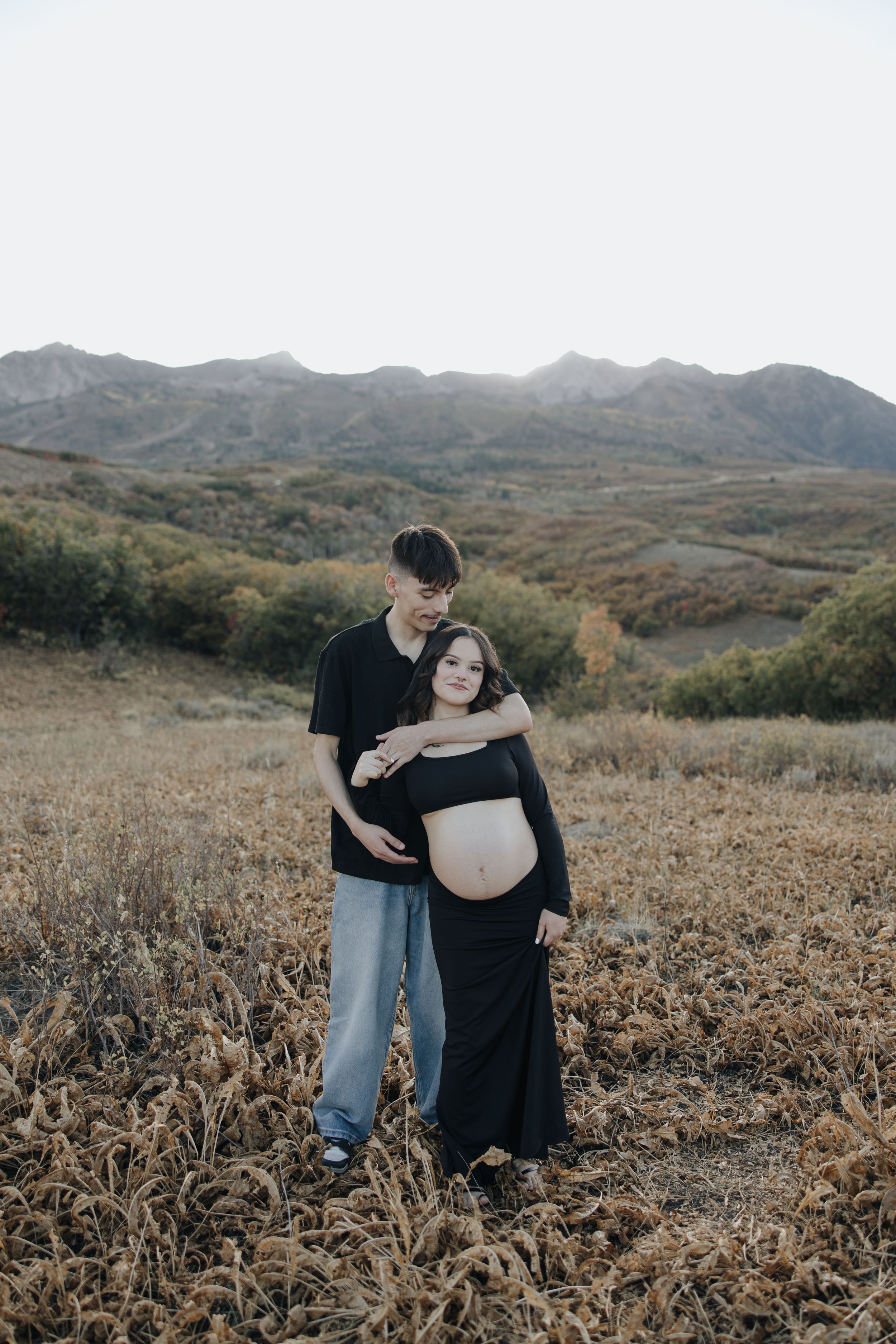 A couple shares an intimate maternity moment together in a golden field with mountains in the background.