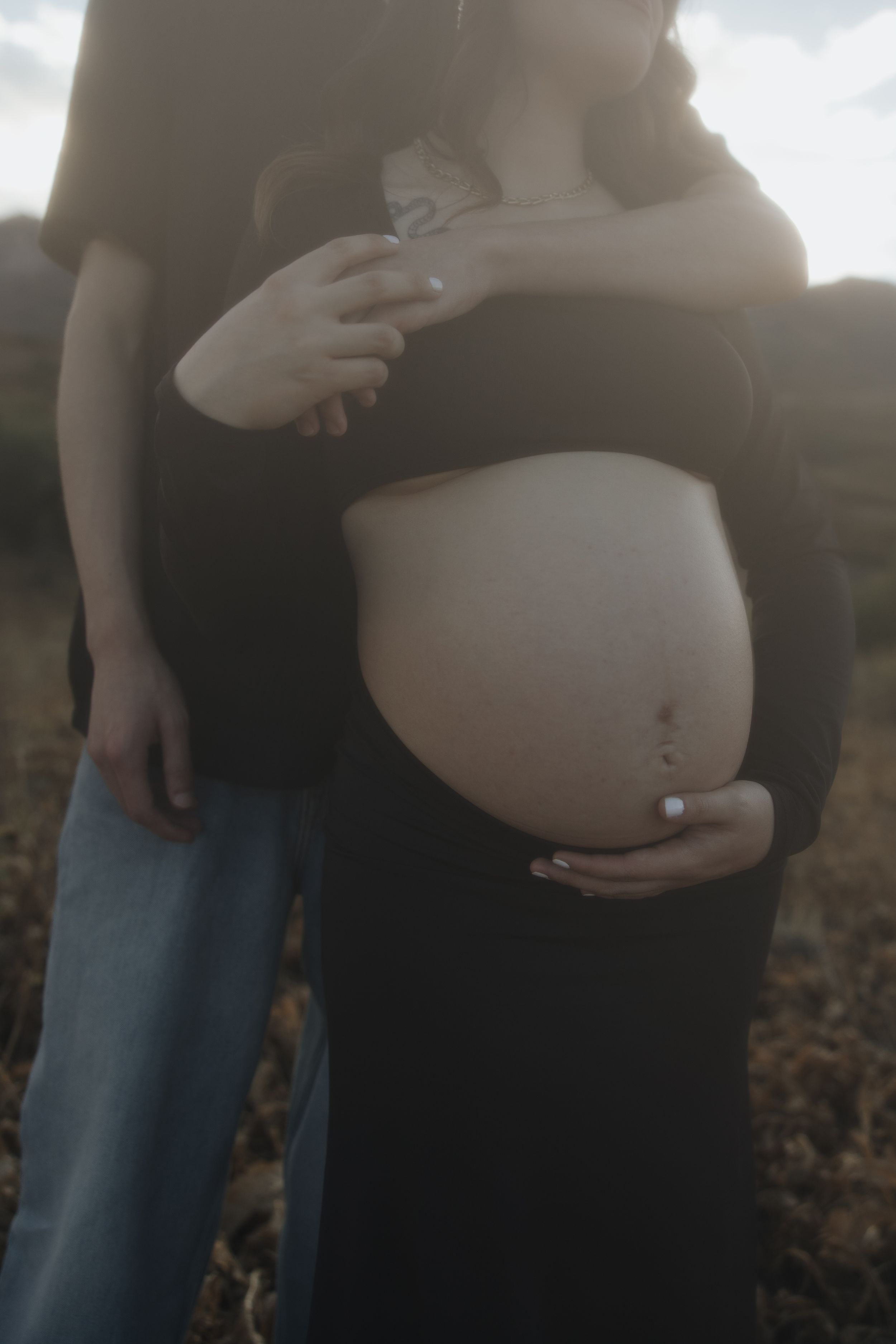 Close up of pregnant belly being cradled in loving hands against dark clothing at dusk.