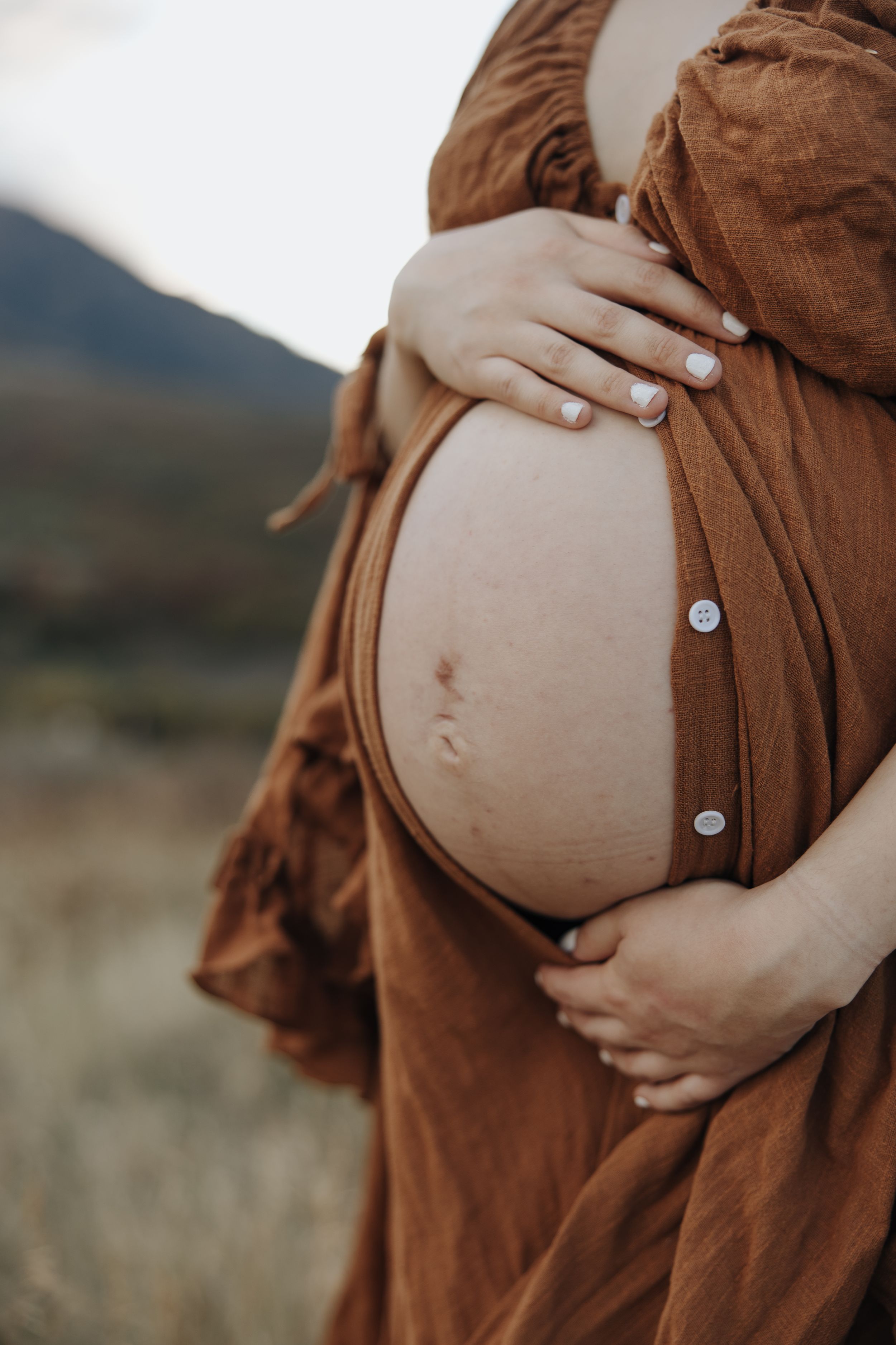 Close up of pregnant belly in brown flowing dress with gentle hands cradling the bump against a mountain landscape.