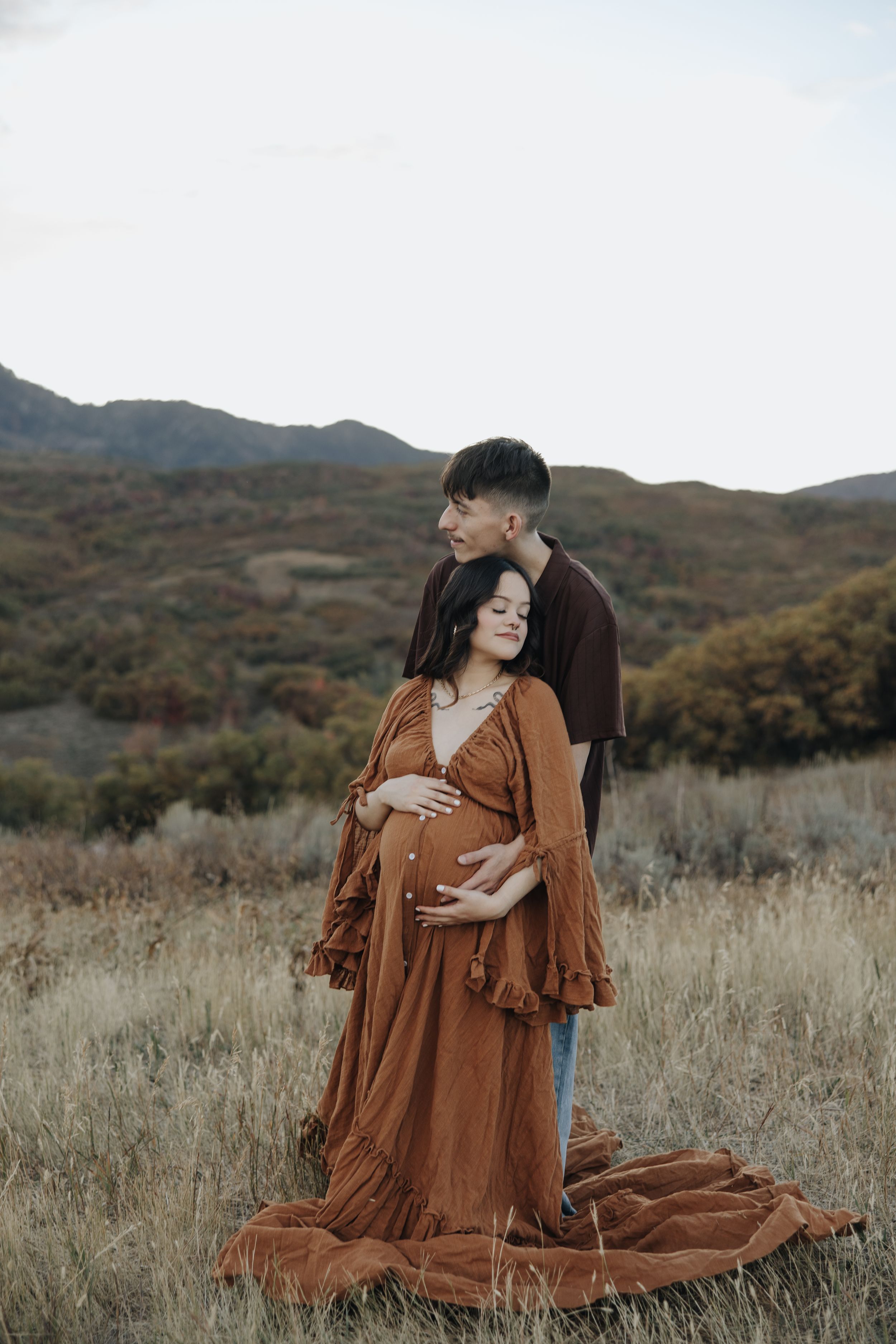 Couple in brown clothing embracing during maternity photoshoot in an autumn mountain meadow.