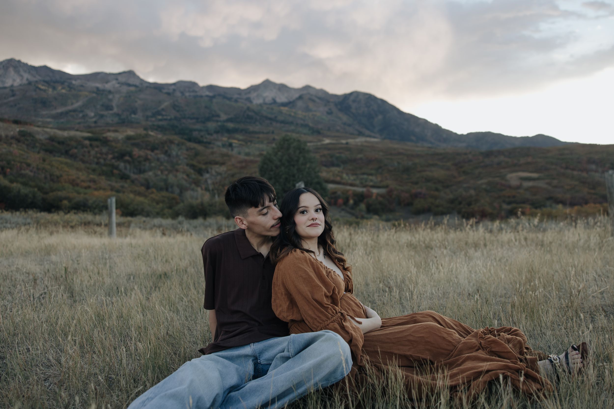 A couple sits together in tall grass with mountains in the backdrop during golden hour.