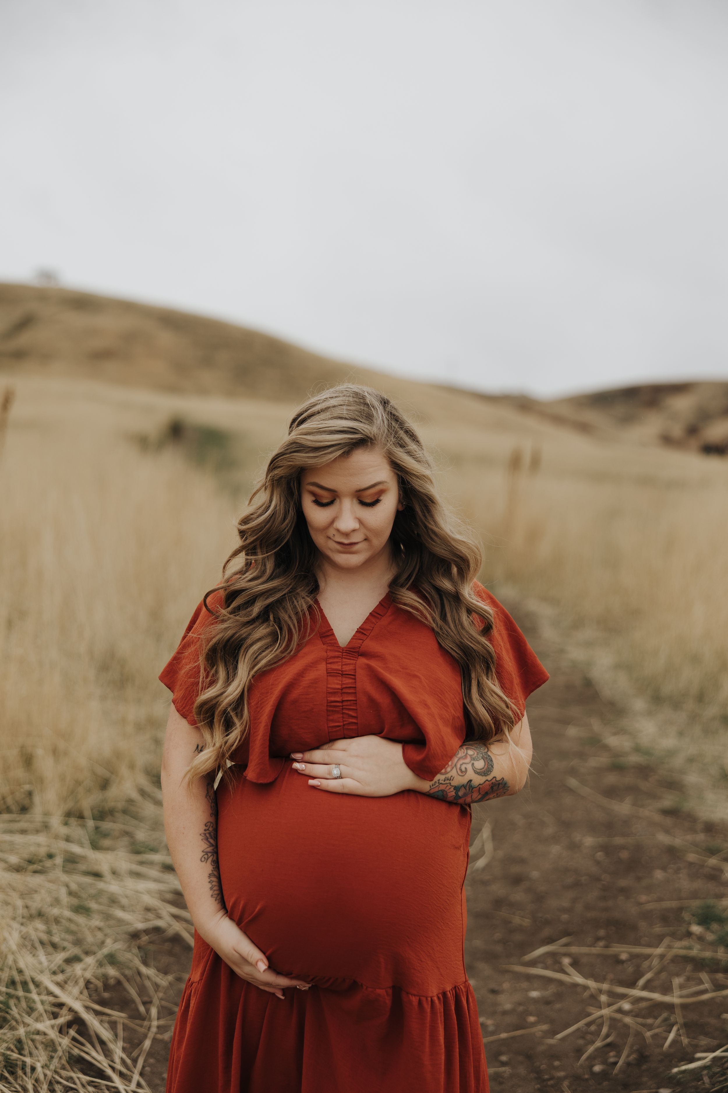 A pregnant woman in a flowing red dress stands in a golden field at sunset, lovingly touching her baby bump.