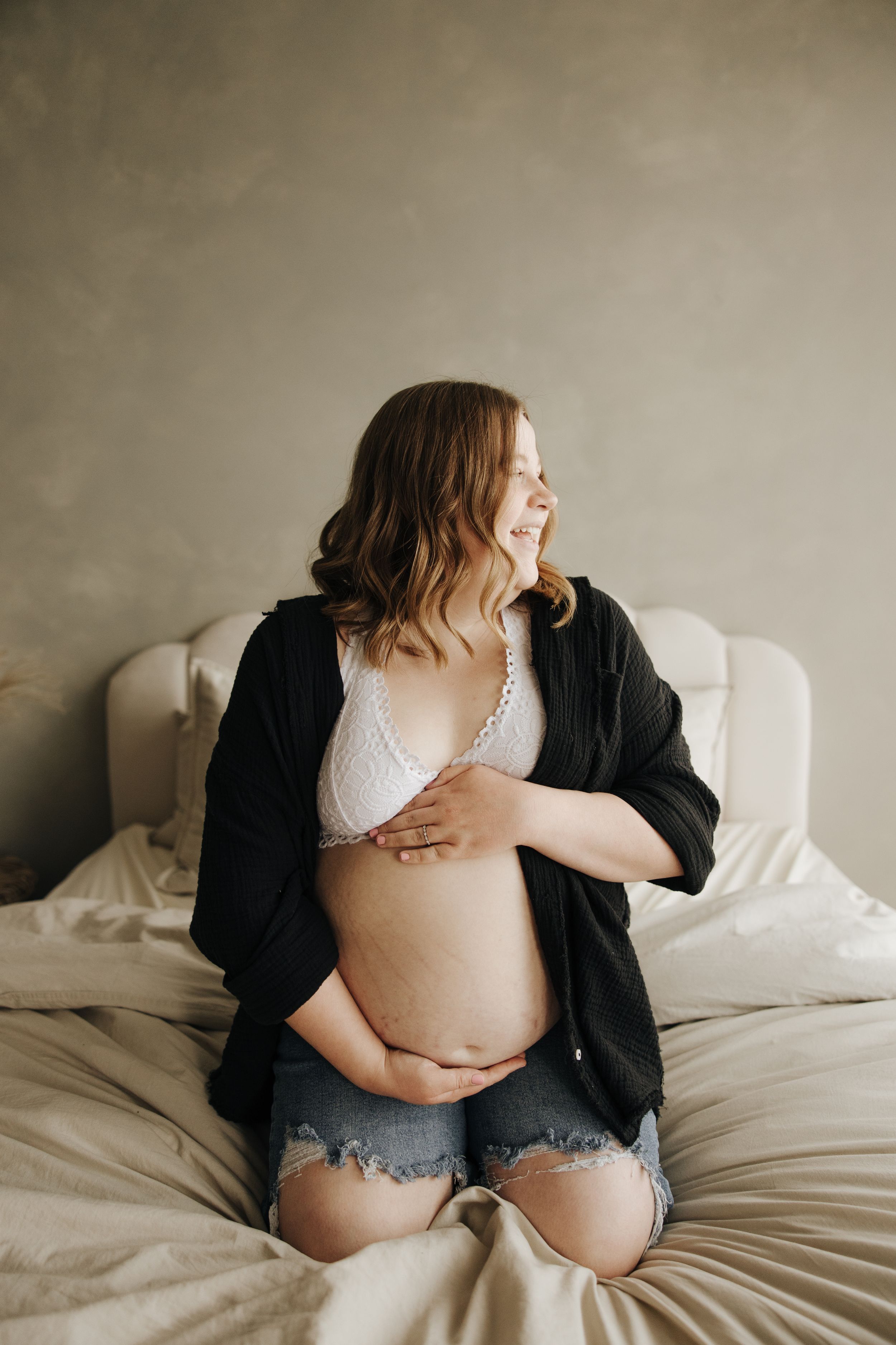 Intimate maternity photo of woman in black cardigan and denim shorts sitting on white bedding.