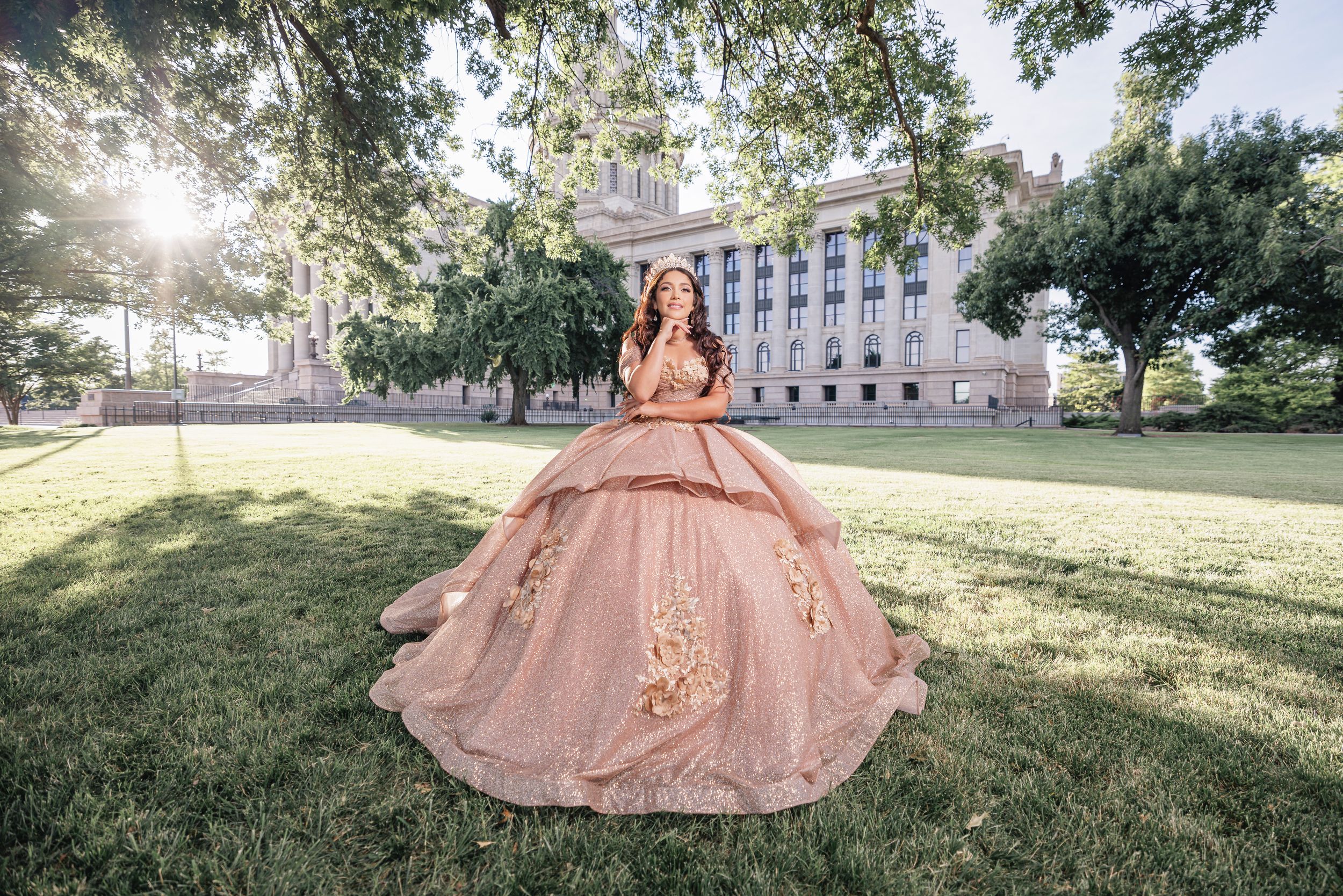 A champagne-colored ball gown photographed on a sunlit lawn with government building backdrop.