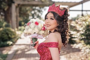 A red off-shoulder gown paired with crown and bouquet in garden setting.