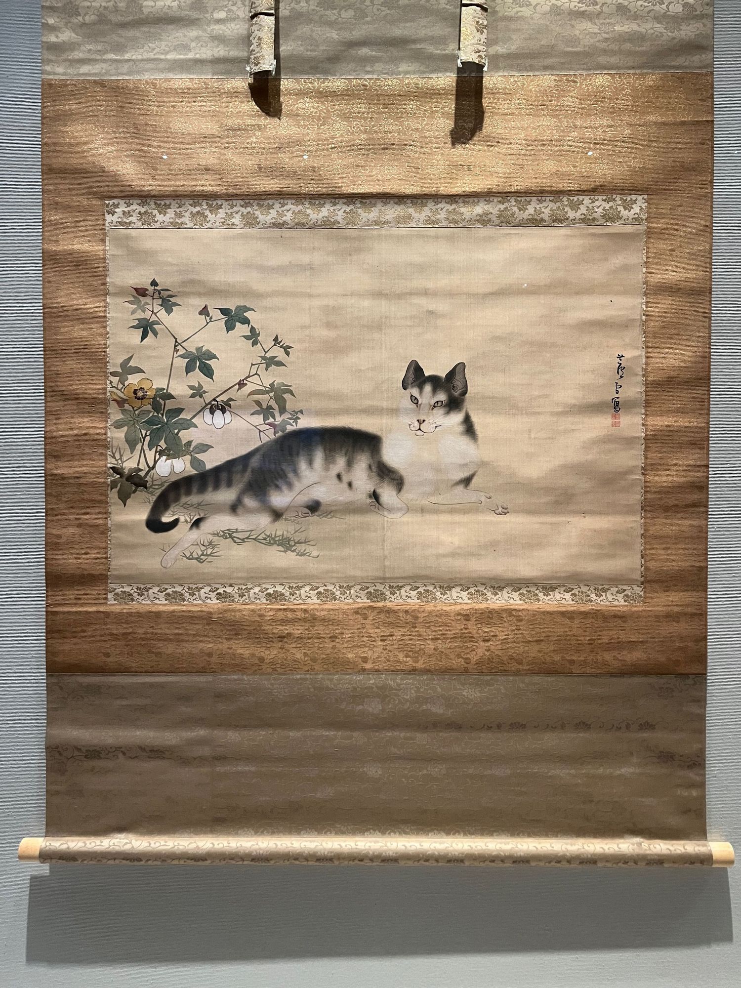 Traditional Japanese scroll painting of a gray and white cat with flowers.