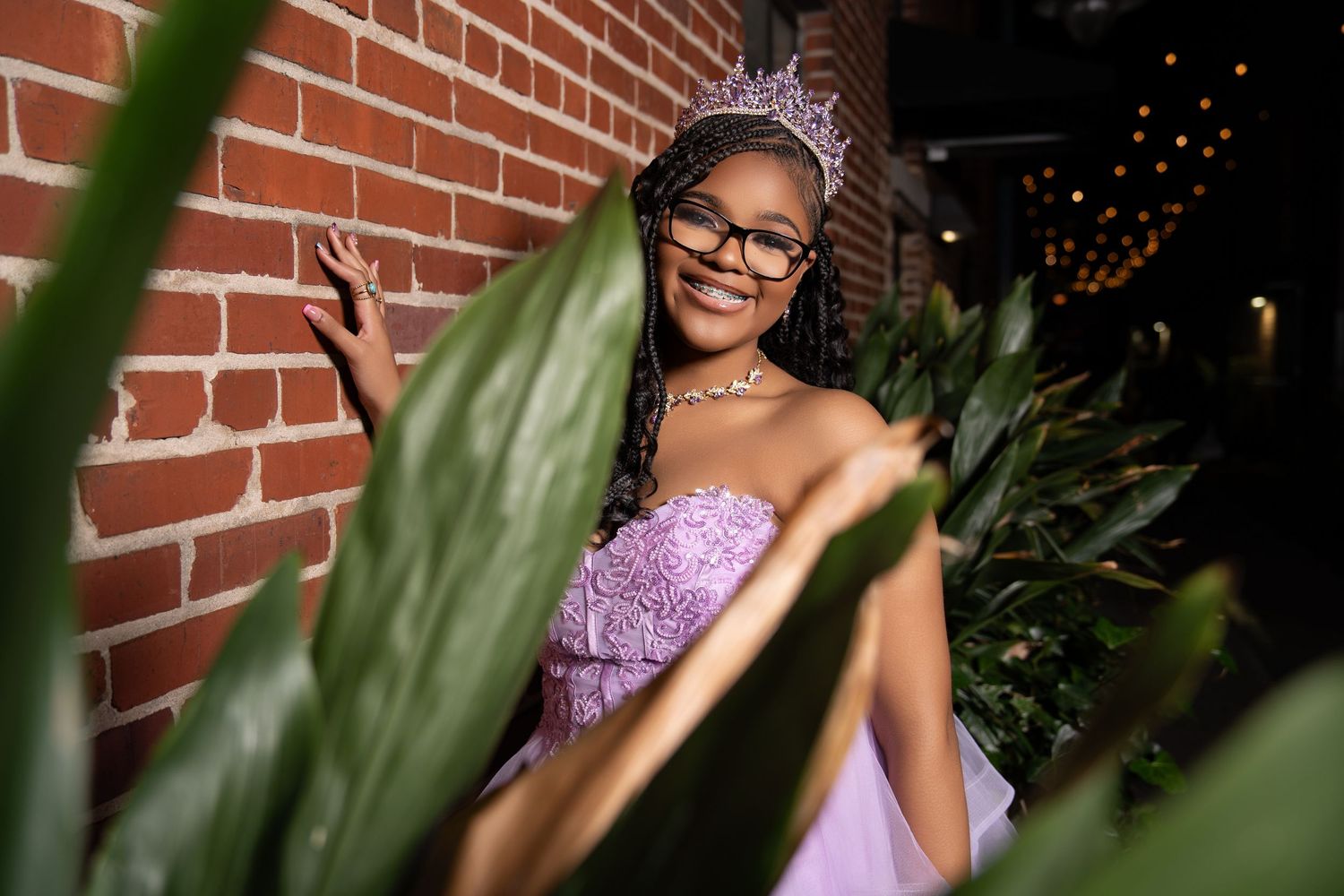 A smiling young woman in glasses, a lavender gown, and a tiara poses beside a red brick wall framed by green plant leaves.