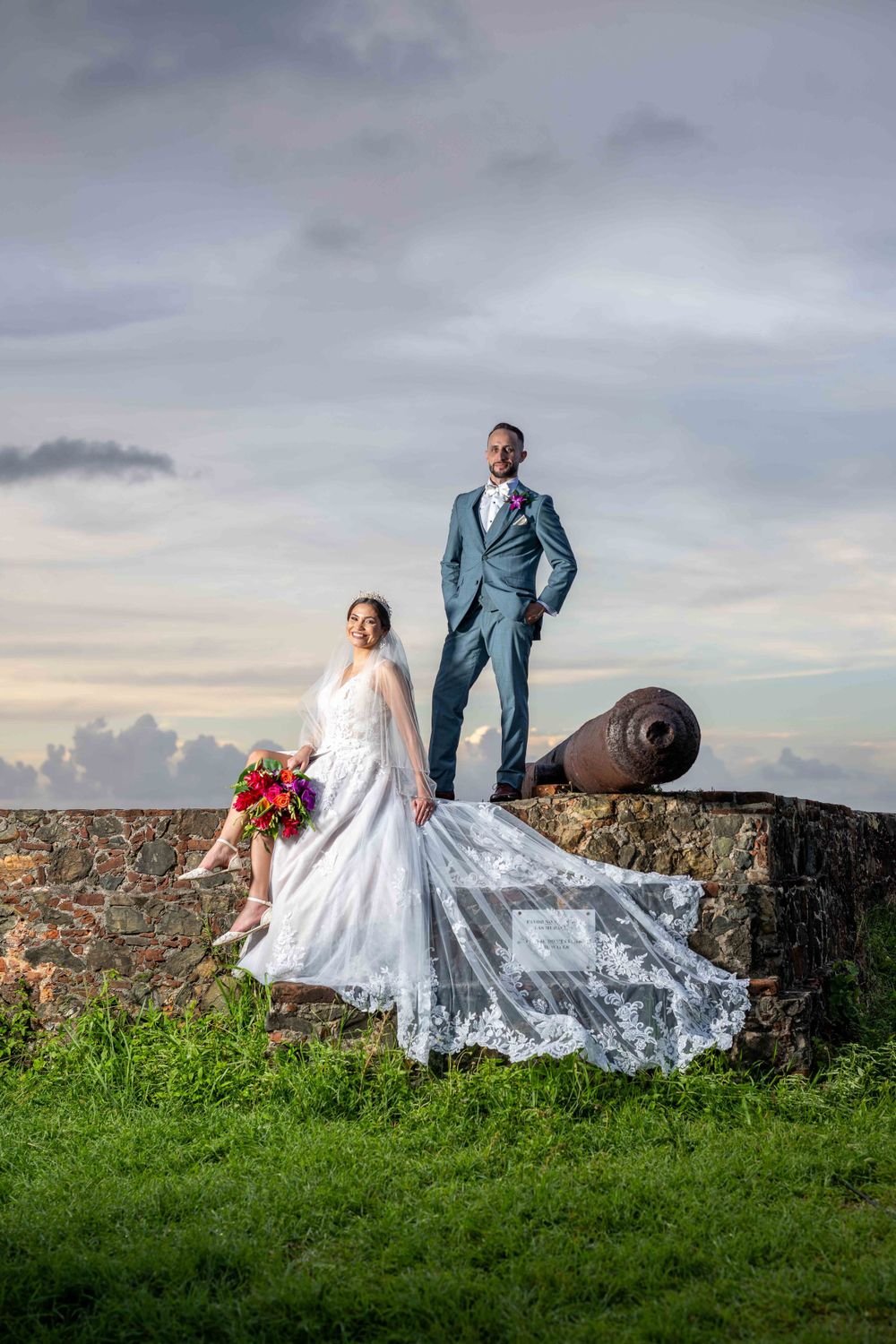 Wedding couple poses dramatically on historic fort wall at sunset with flowing veil.