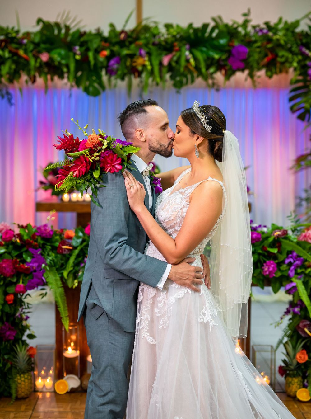 Couple shares romantic kiss under floral arch with purple lighting at wedding reception.