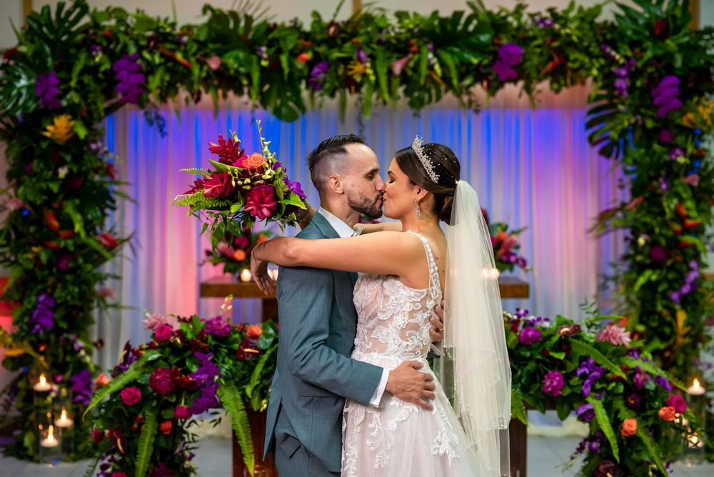 Wedding couple embraces for kiss under elaborate tropical floral arch with purple lighting.