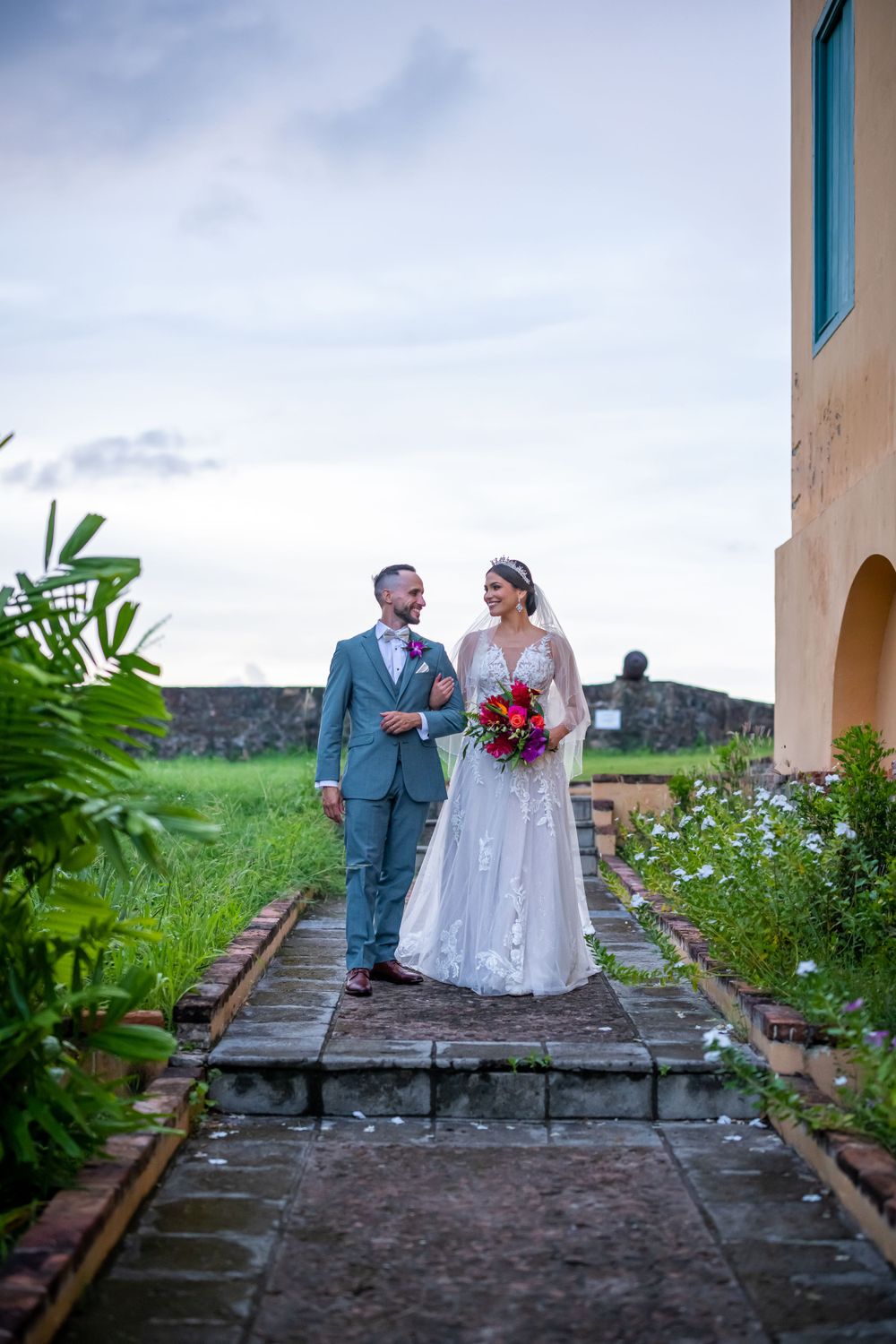 A wedding couple standing on a stone pathway with lush greenery and architecture in the background.