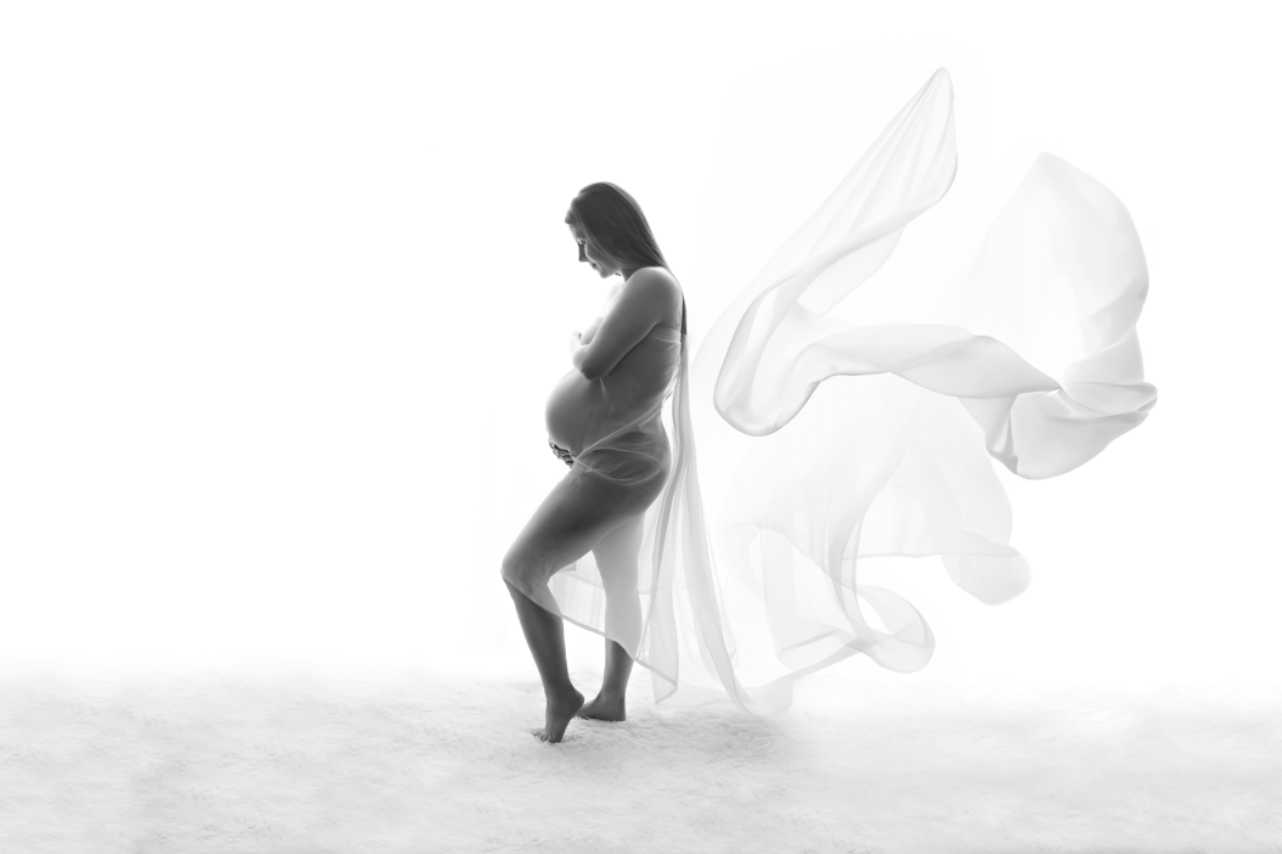 Black and white maternity photo of pregnant woman draped in flowing white fabric against white background. Embracing Life’s Beauty