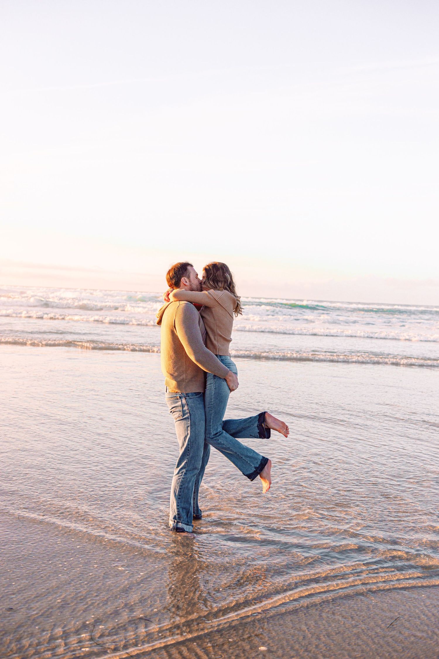 A romantic couple embrace and kiss while standing in shallow ocean waves at sunset on a beach.