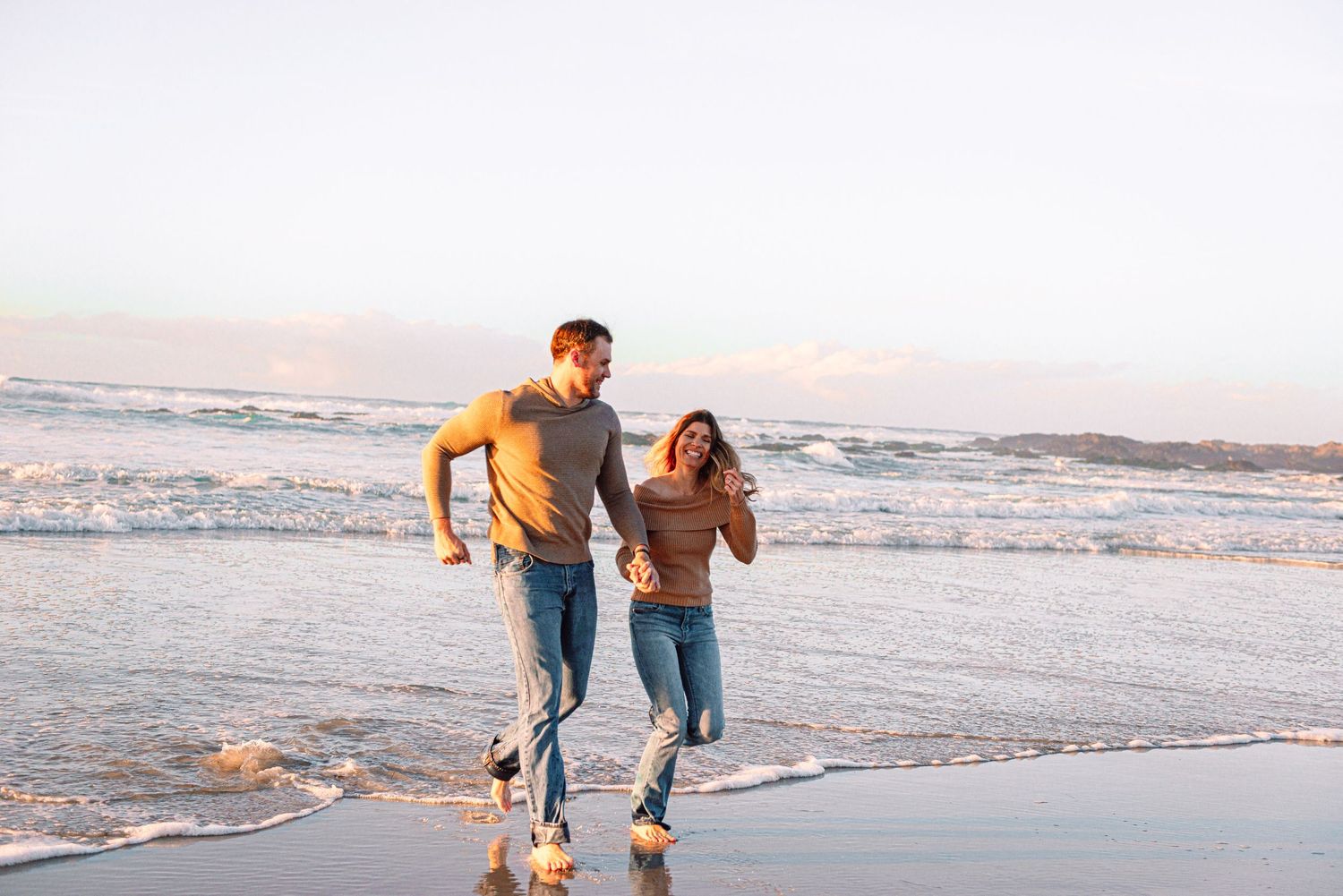 Couple running joyfully together through shallow waves on a beach at dusk.