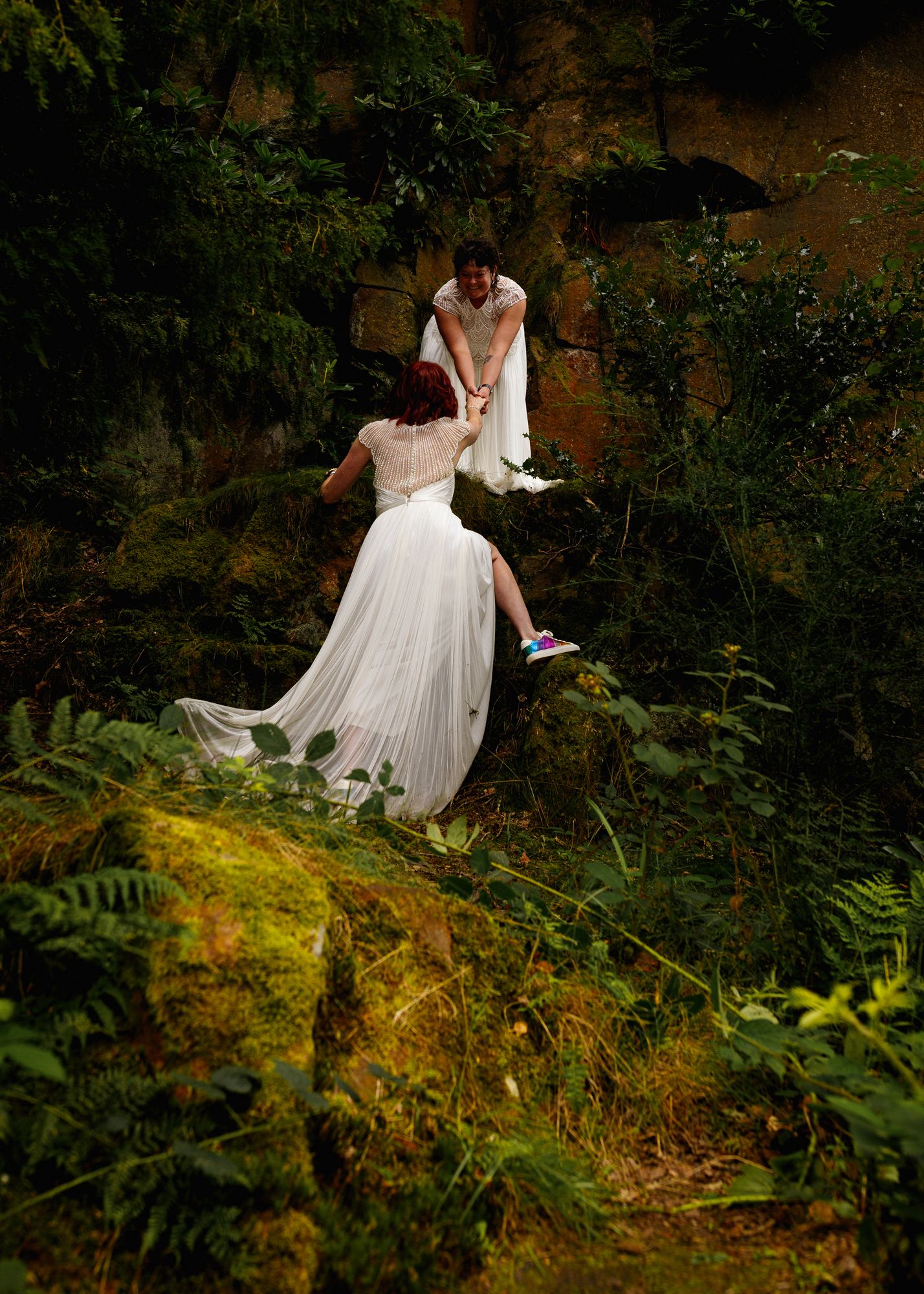 Two people in flowing white dresses explore a mossy forest cliff covered in lush greenery and ferns.