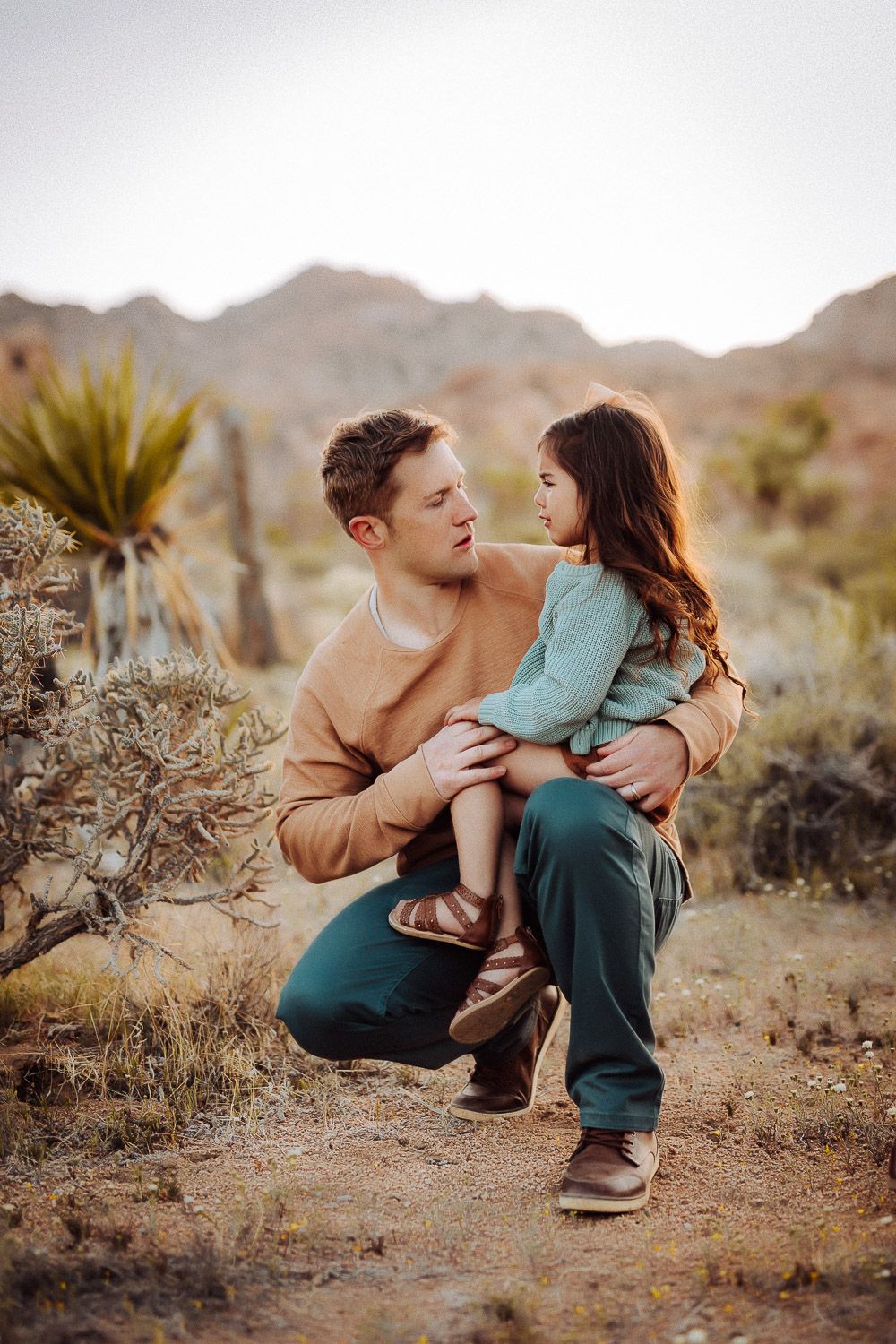 Father embracing his child in the Joshua Tree desert, surrounded by natural scenery and mountains at sunset.