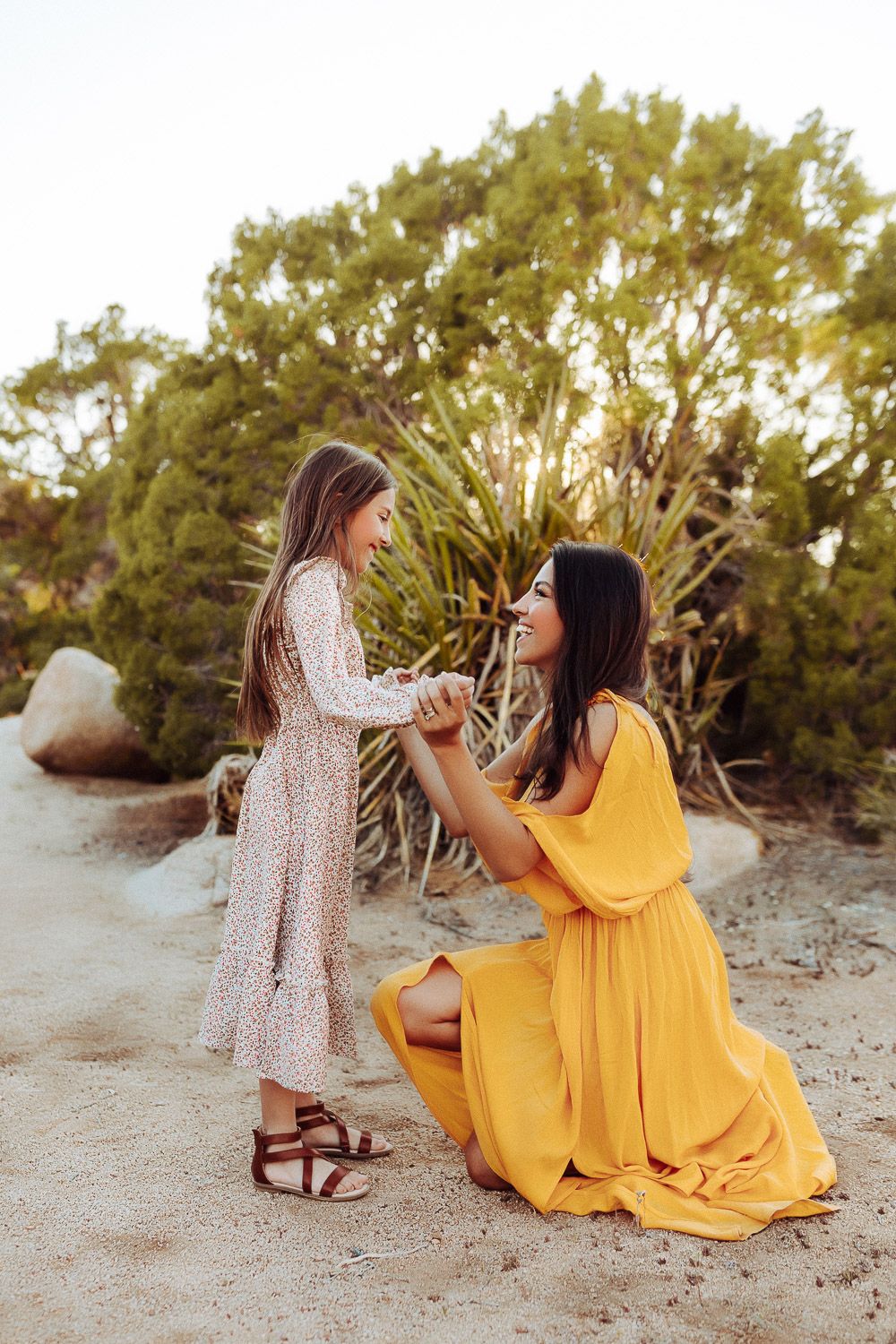 “Mother and daughter holding hands in the Joshua Tree desert, sharing a tender moment with a scenic mountain backdrop.