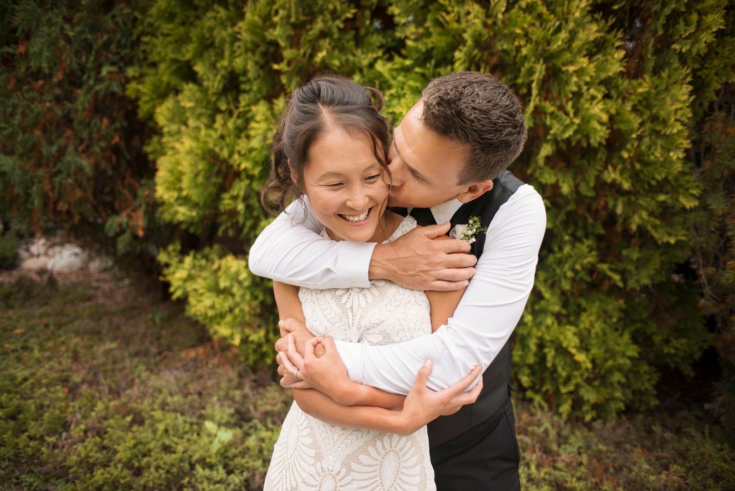 Groom in suit hugging bride in wedding dress  and kissing cheek. Romantic, authentic, classic wedding photo. Greenery. Beautiful lighting.