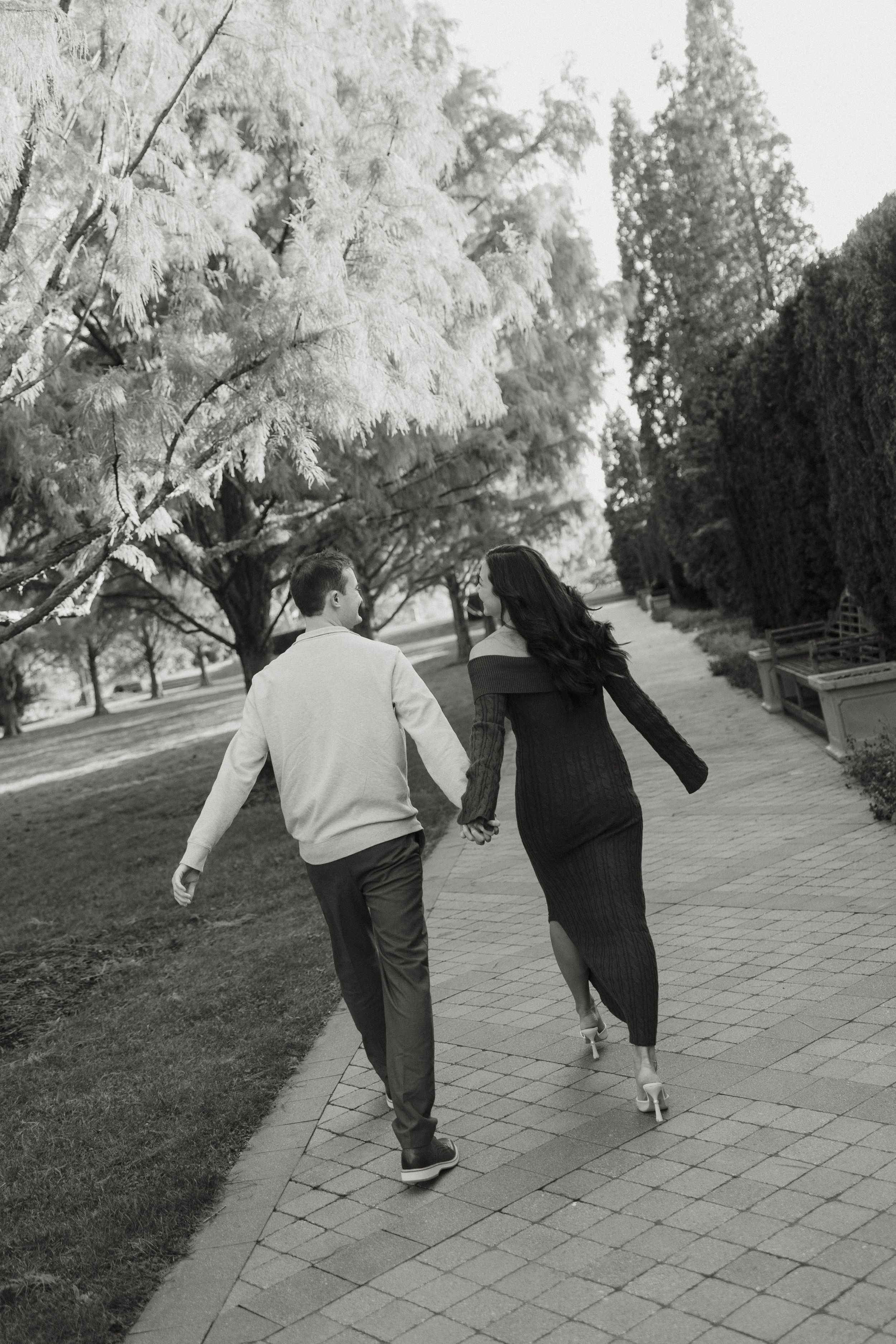 A couple walking together hand in hand along a tree-lined brick path in black and white photography.