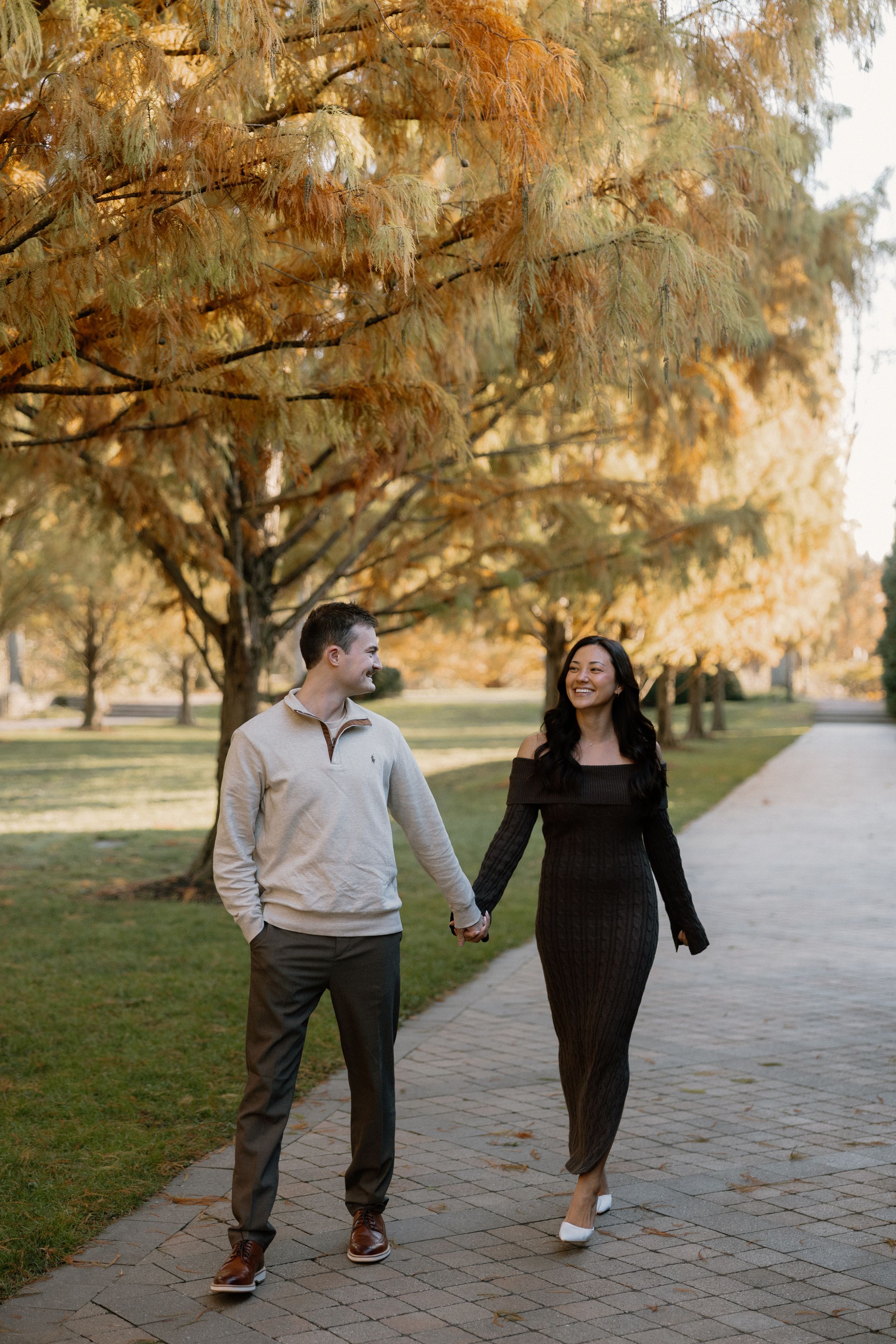 A couple walks together on a paved path surrounded by autumn trees with golden foliage.