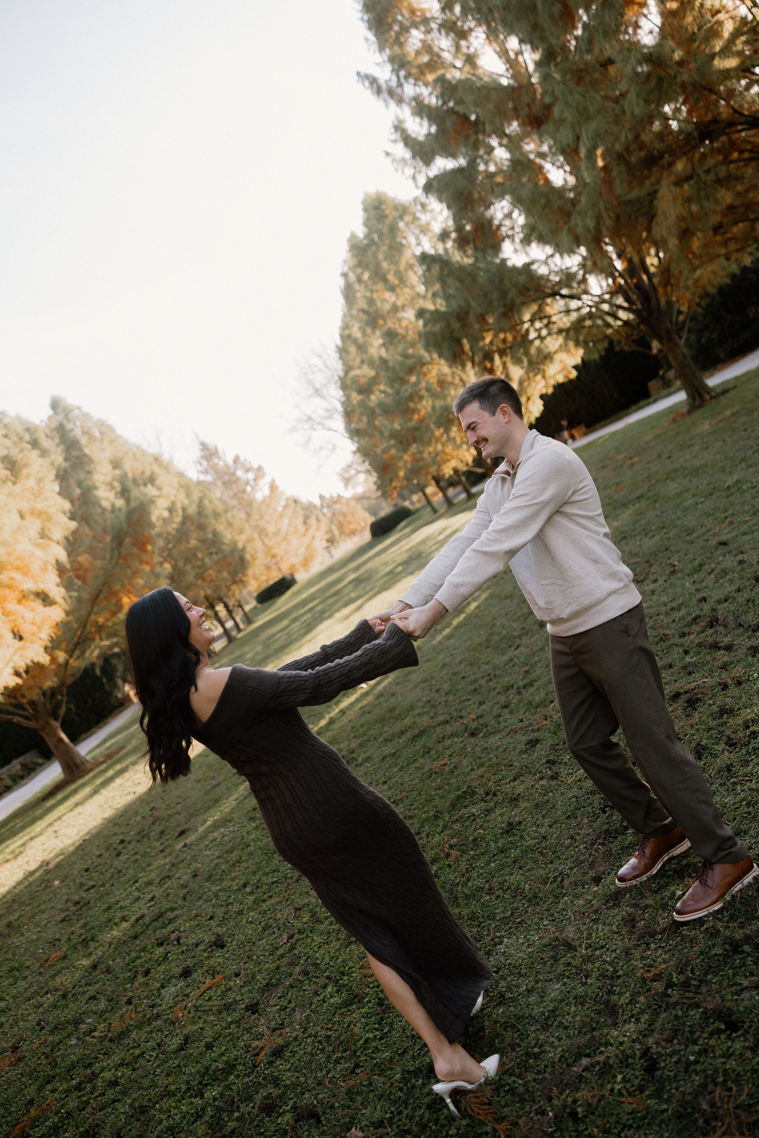 A couple playfully falls down a grassy hillside at sunset with tall trees in the background.